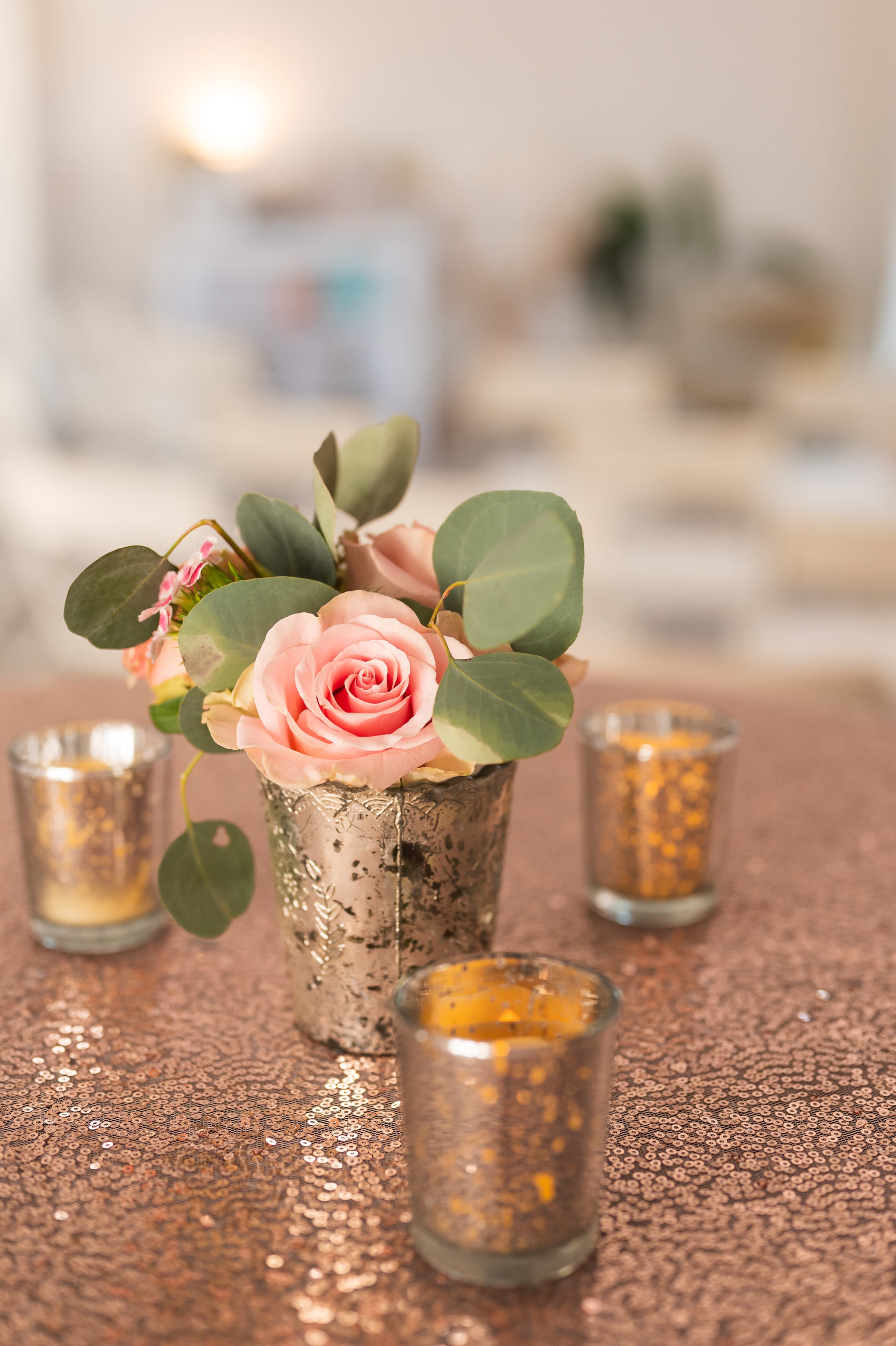 A silver-painted pot holds pink roses and green leaves, surrounded by three small glass votive candles, on a glittering copper tablecloth.