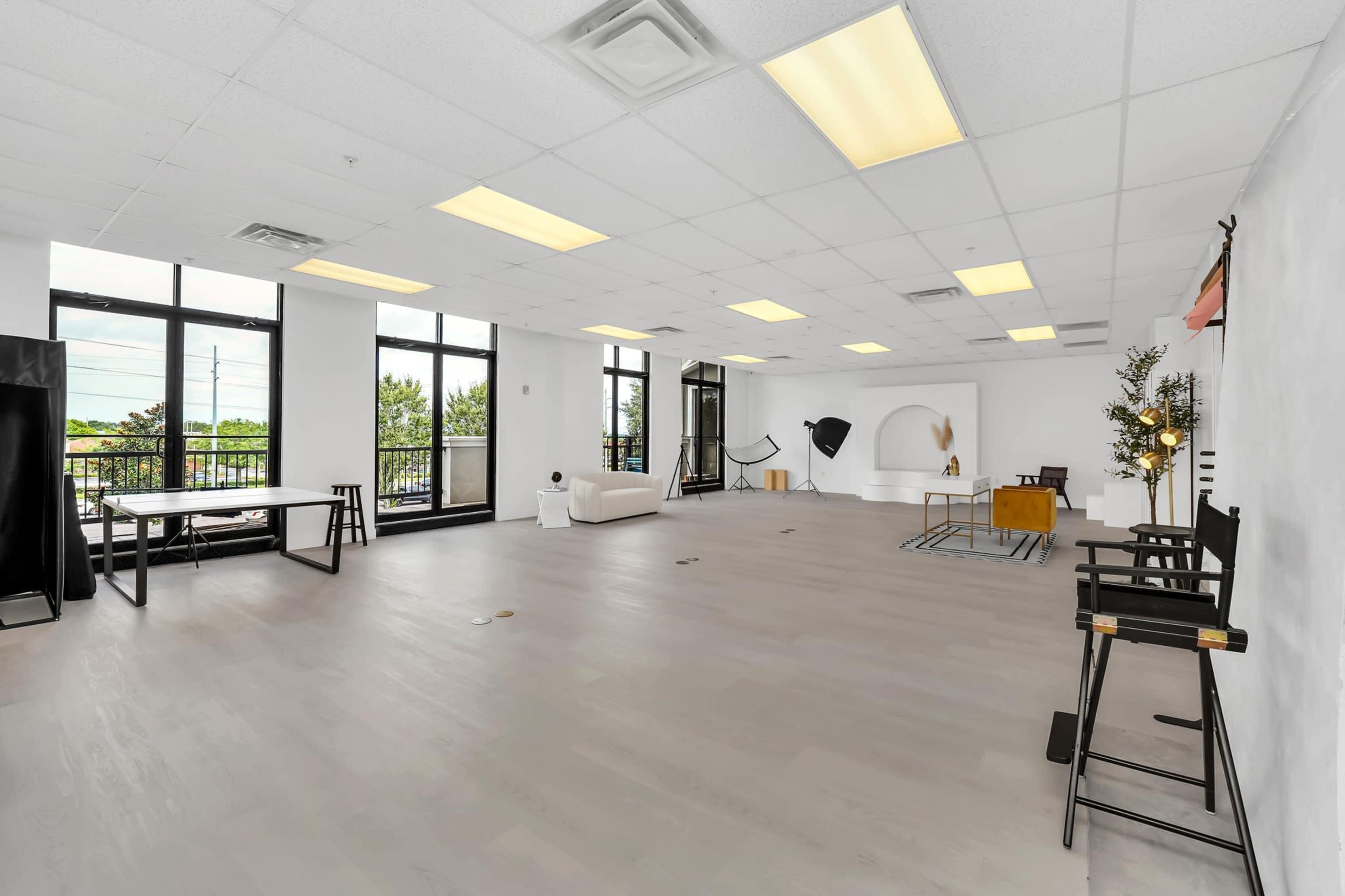 The image shows a spacious, well-lit room with large windows, featuring minimal furniture like a white couch, a table, and several empty chairs.