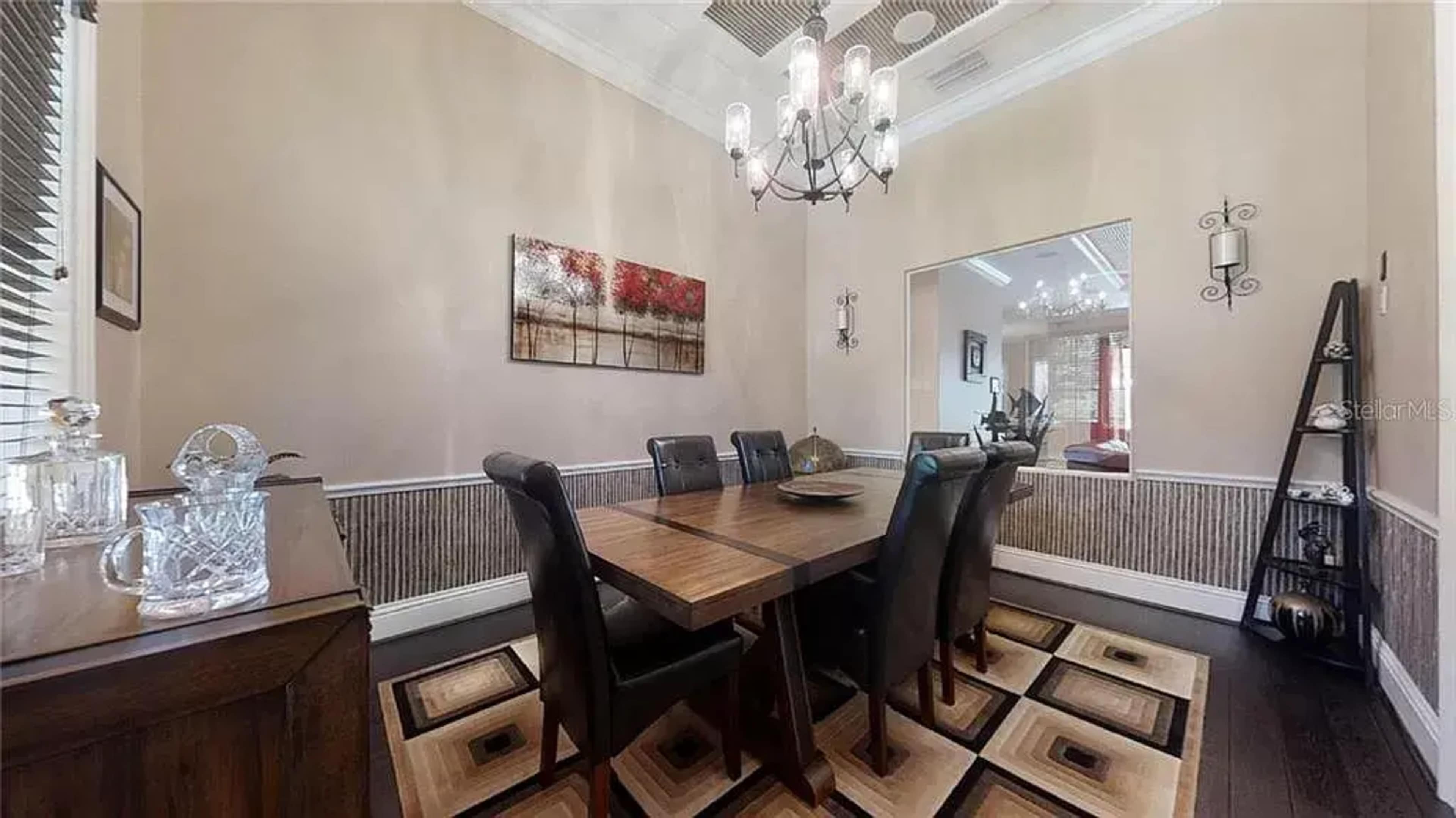 A dining room features a wooden table surrounded by six black chairs, with a chandelier overhead and a large artwork on the wall.