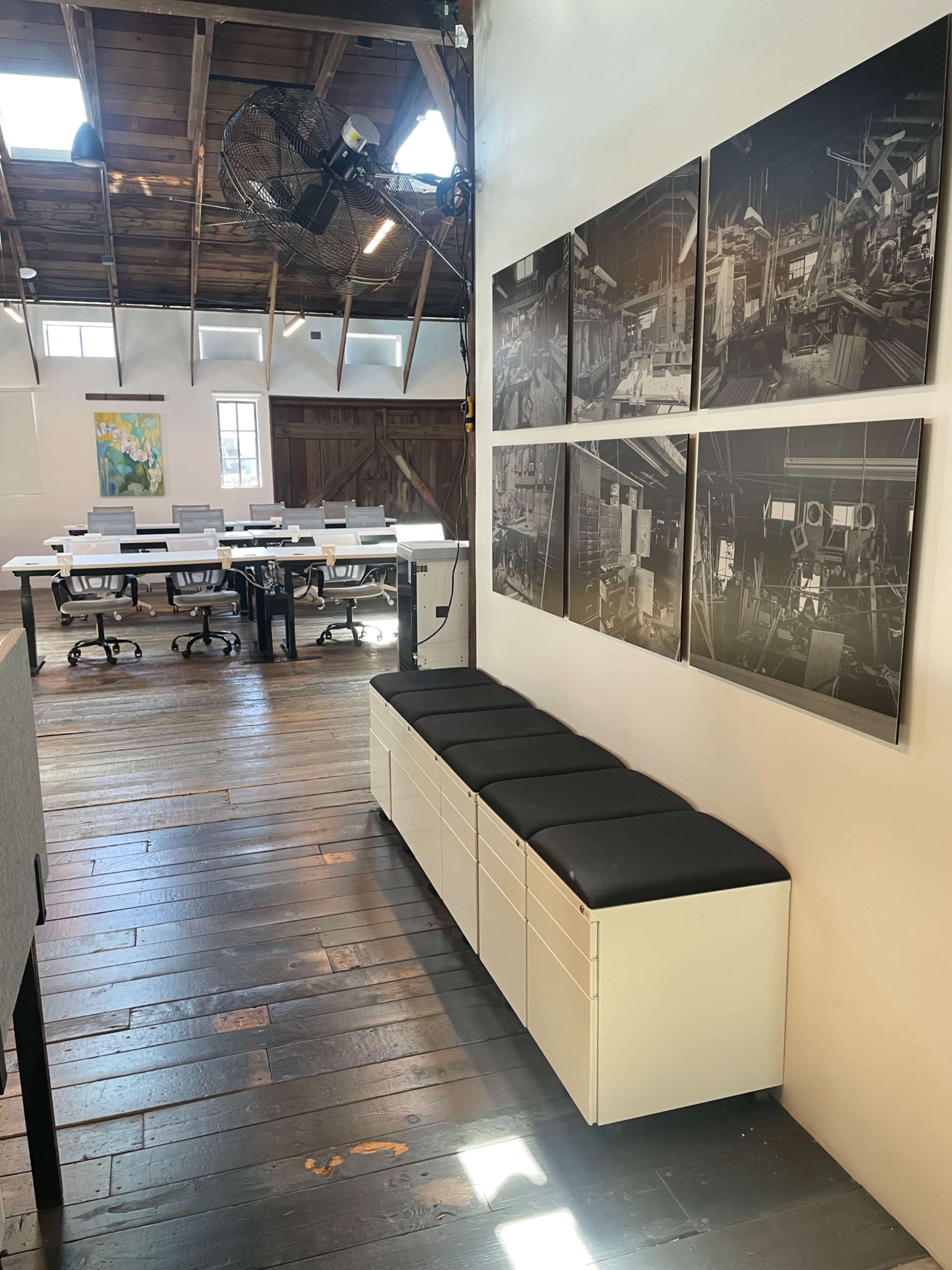 The image shows a modern office space with a seating area along one wall and several black-and-white photographs displayed beside it, while a row of desks with computers is visible in the background.