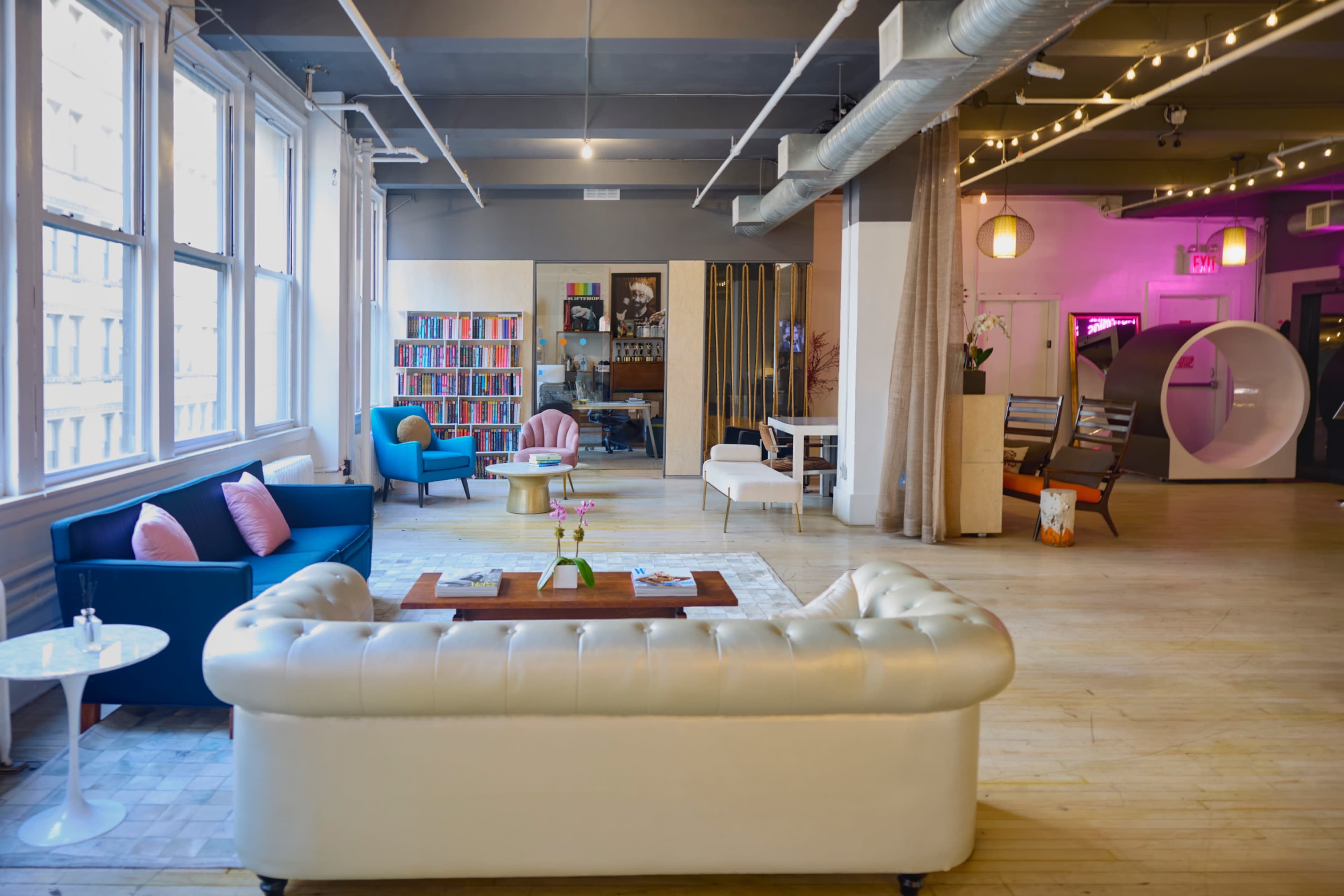 The image shows a spacious, modern living area with a mix of colorful chairs, a white tufted sofa, bookshelves, and ambient lighting.