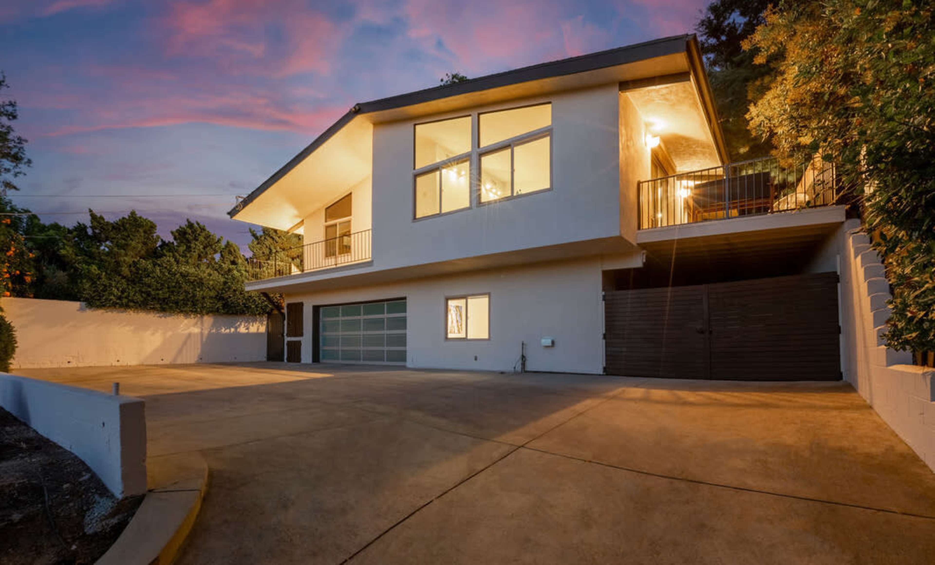 A modern two-story house with large windows and a balcony sits on a driveway at dusk.