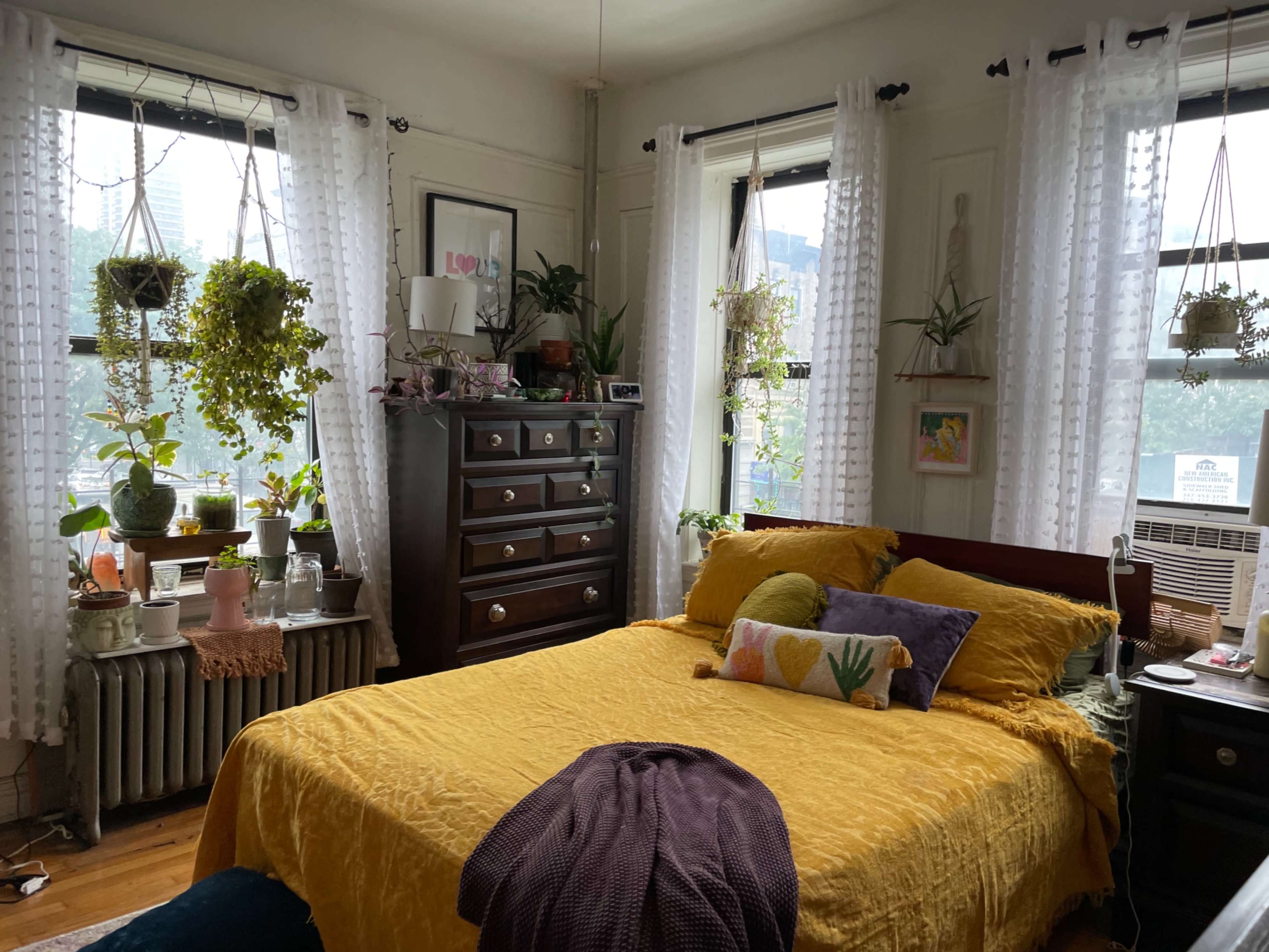 A cozy bedroom featuring a yellow bedspread, multiple plants on window sills, and a large wooden dresser with decorative items.