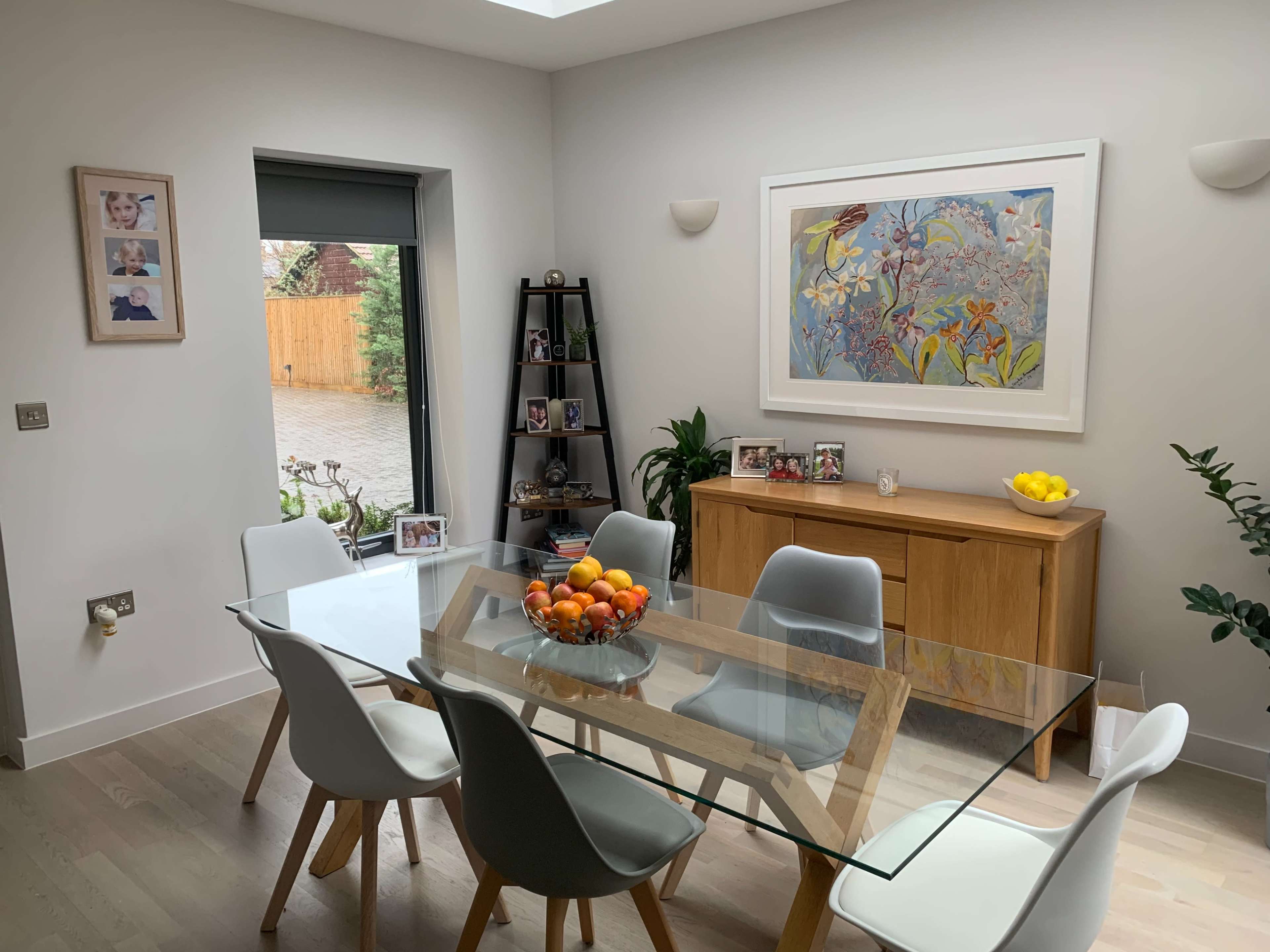 A dining area features a glass table surrounded by white chairs, with a wooden sideboard, framed artwork, and a bowl of fruit centerpiece.