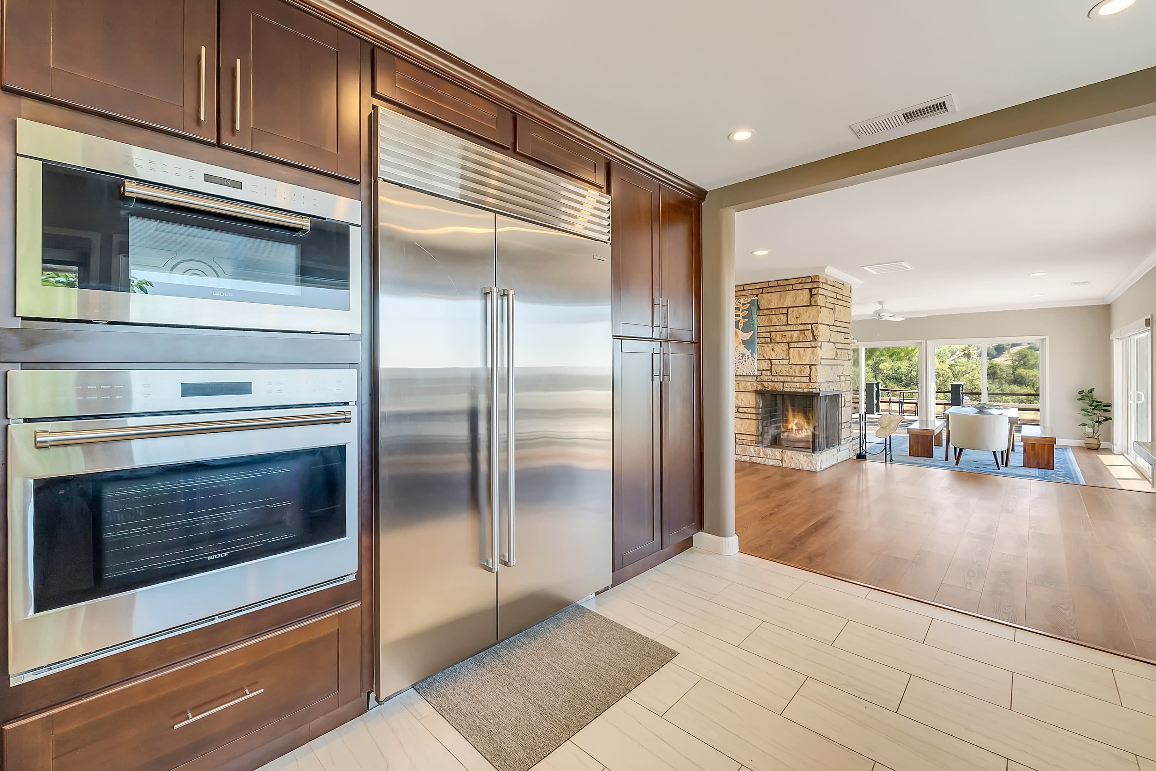 A modern kitchen with stainless steel appliances, including a refrigerator and double ovens, opening up to a living area with a stone fireplace and large windows.