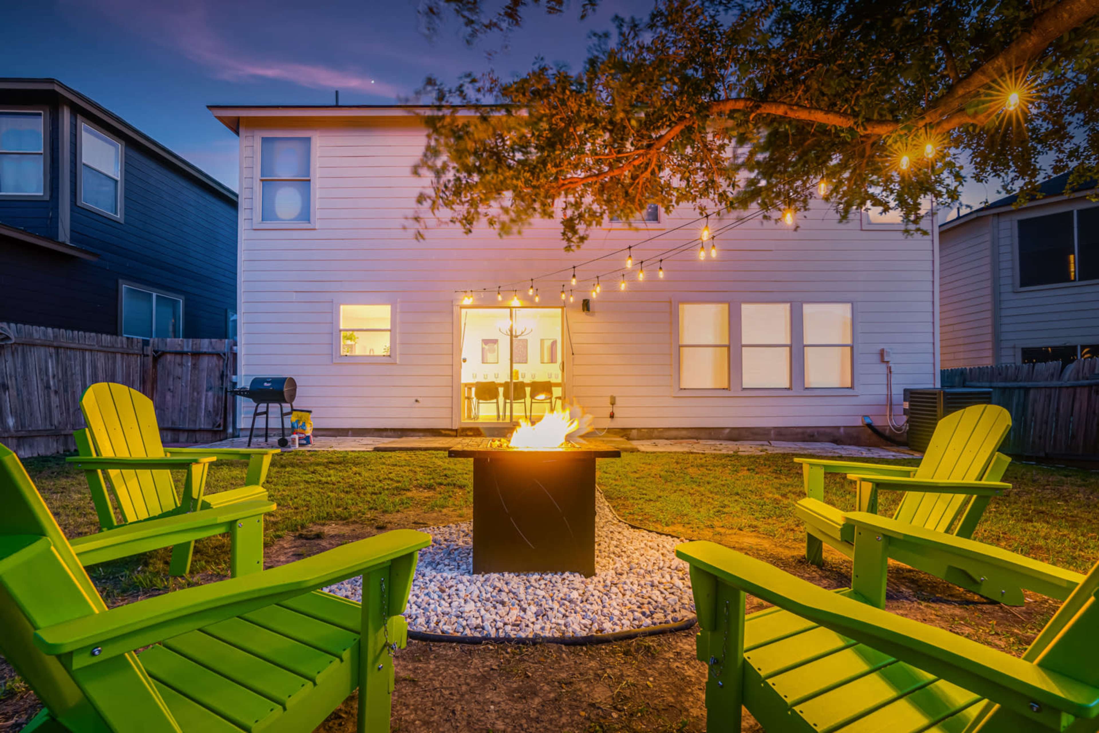 A backyard scene features a fire pit surrounded by four green chairs, with a house and string lights in the background.