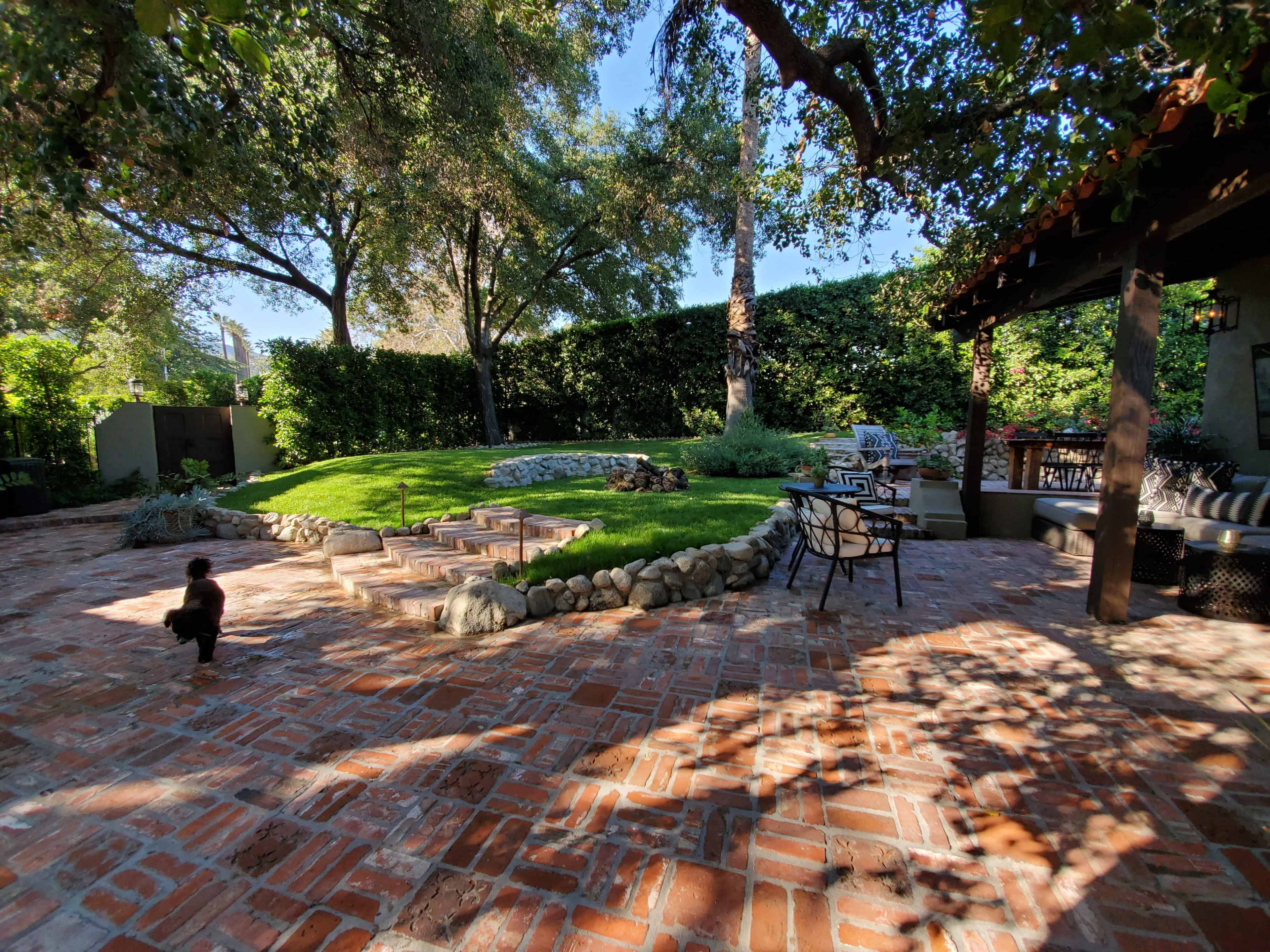The image shows a landscaped backyard with a stone pathway, grass area, and seating arrangements under a shaded patio.