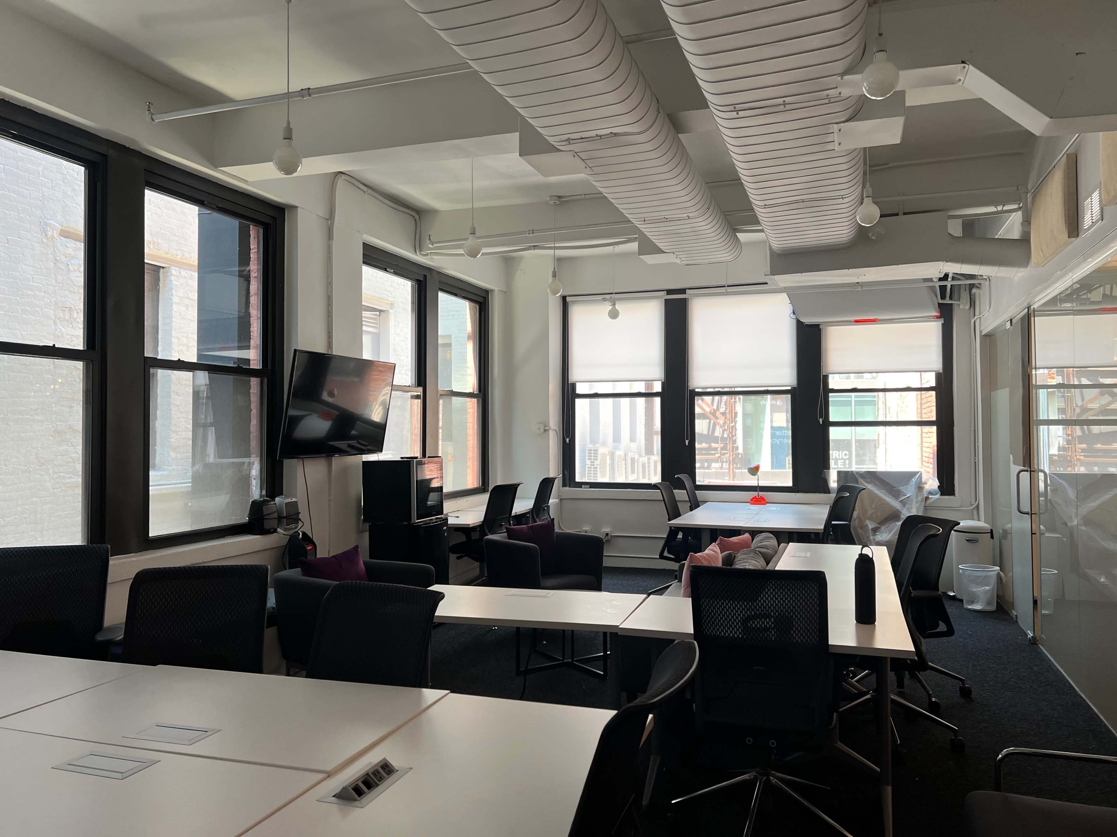 The image shows an office space with large windows, several desks arranged in the room, and a television mounted on the wall.