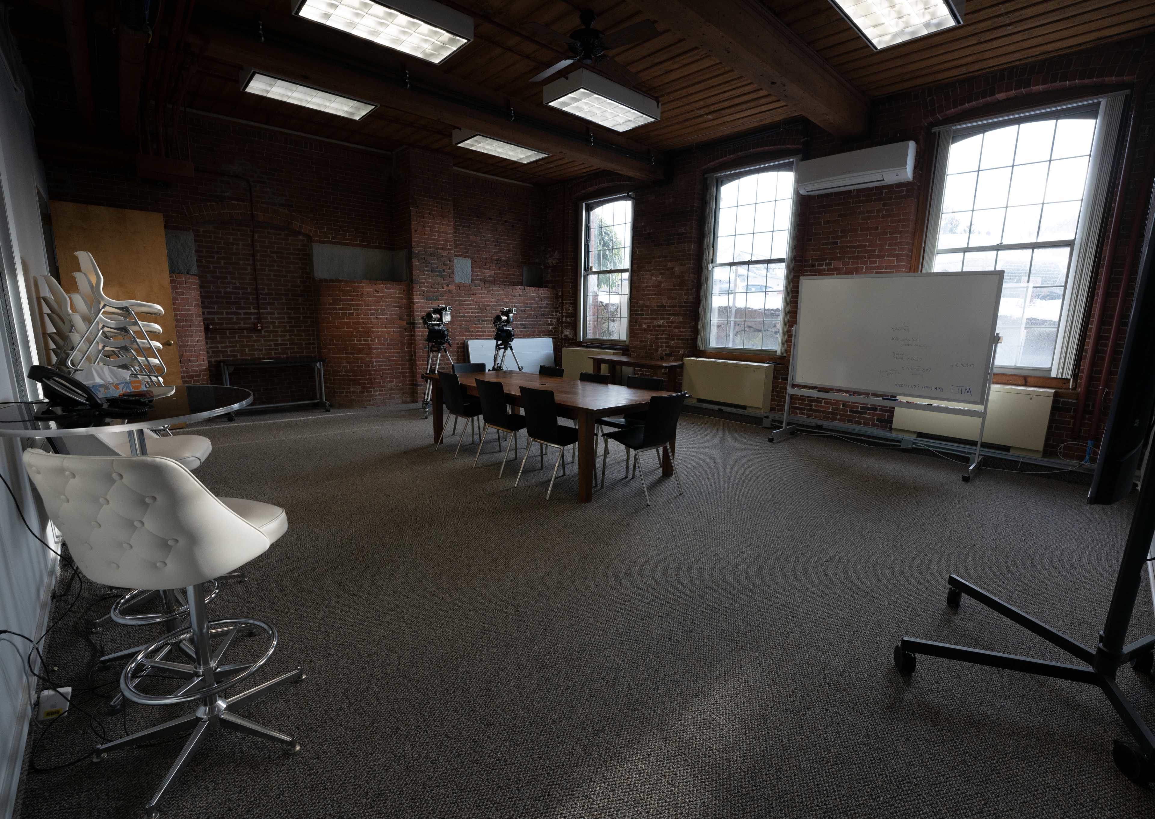 A spacious meeting room with a long table, several chairs, and a whiteboard, surrounded by brick walls and large windows.