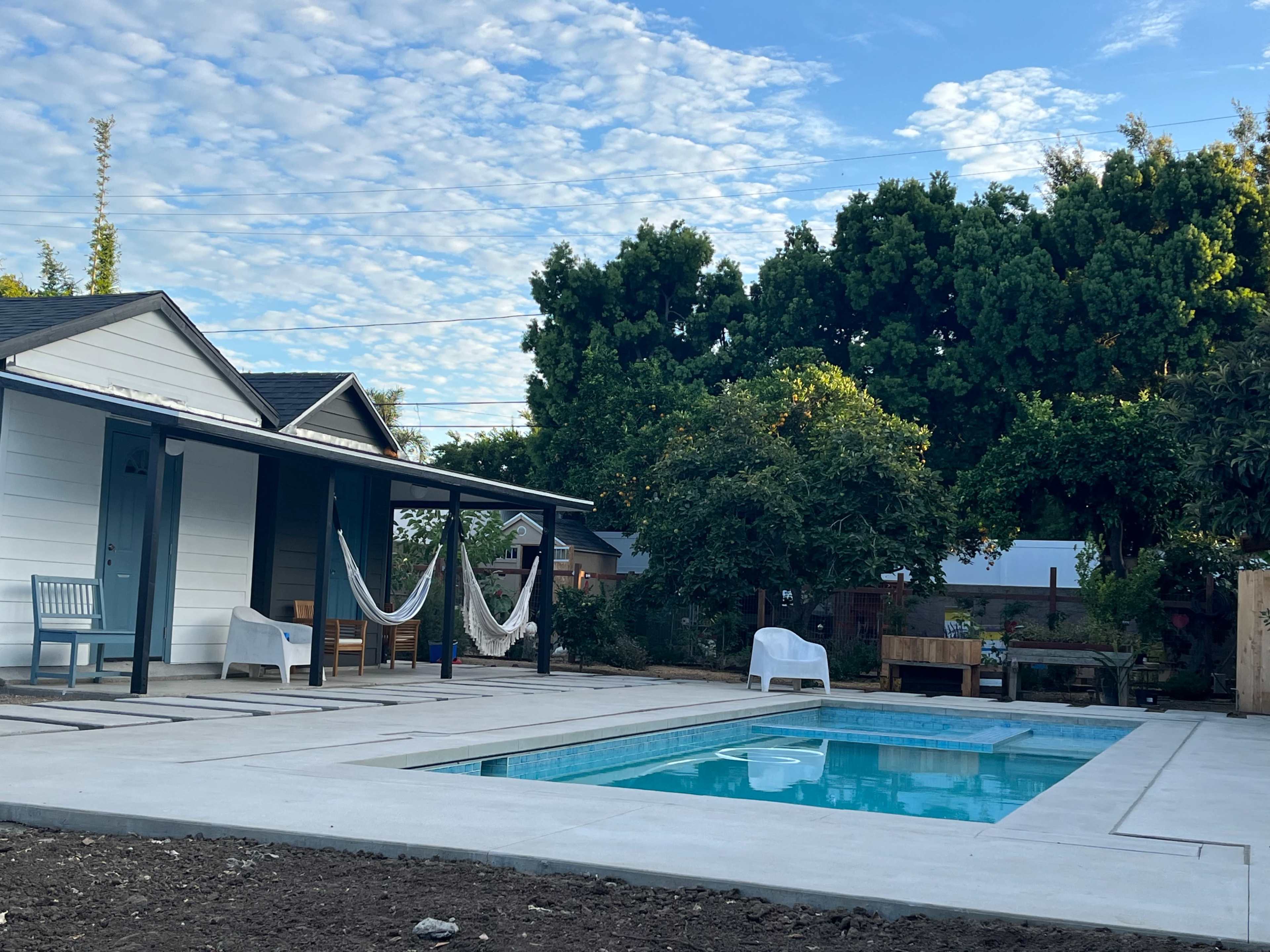 A rectangular swimming pool is situated between two houses, with hammocks hanging nearby and a garden area in the background.