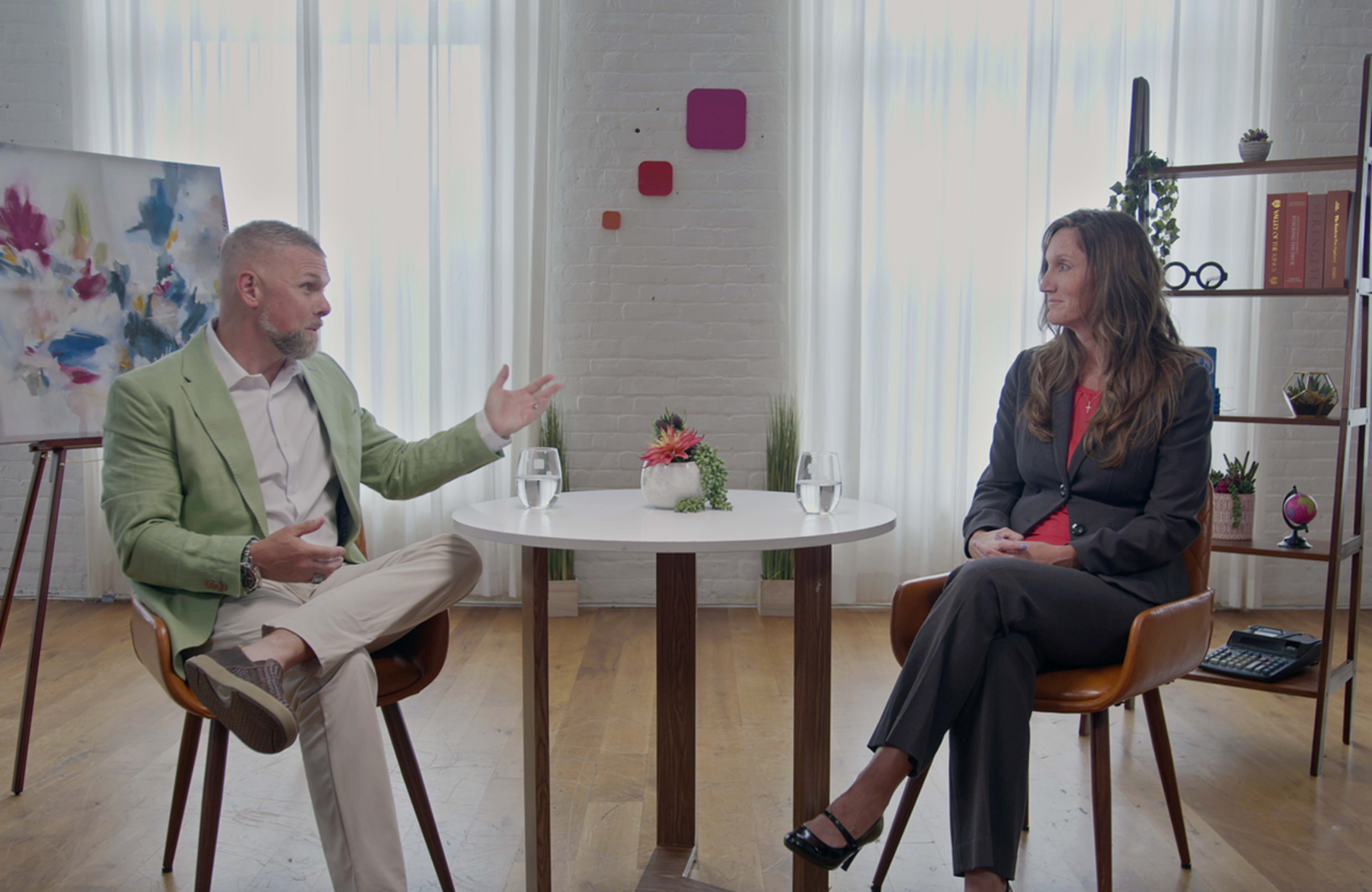A man in a light green blazer and a woman in a dark suit sit at a small round table with water glasses, discussing in a well-lit, minimalist room.