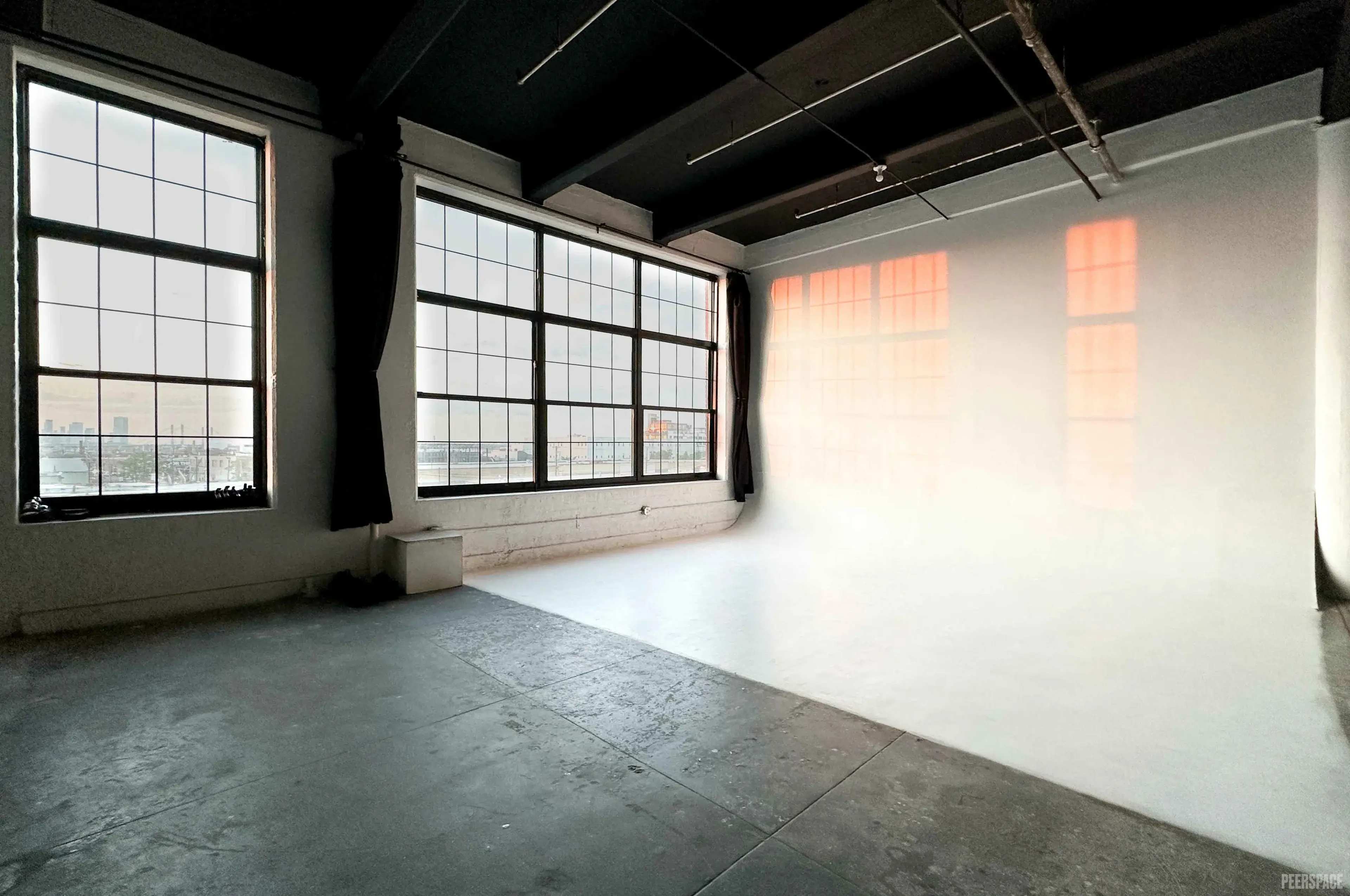 The image shows a spacious, well-lit interior with large windows and a plain white wall, creating an open and minimalistic space.