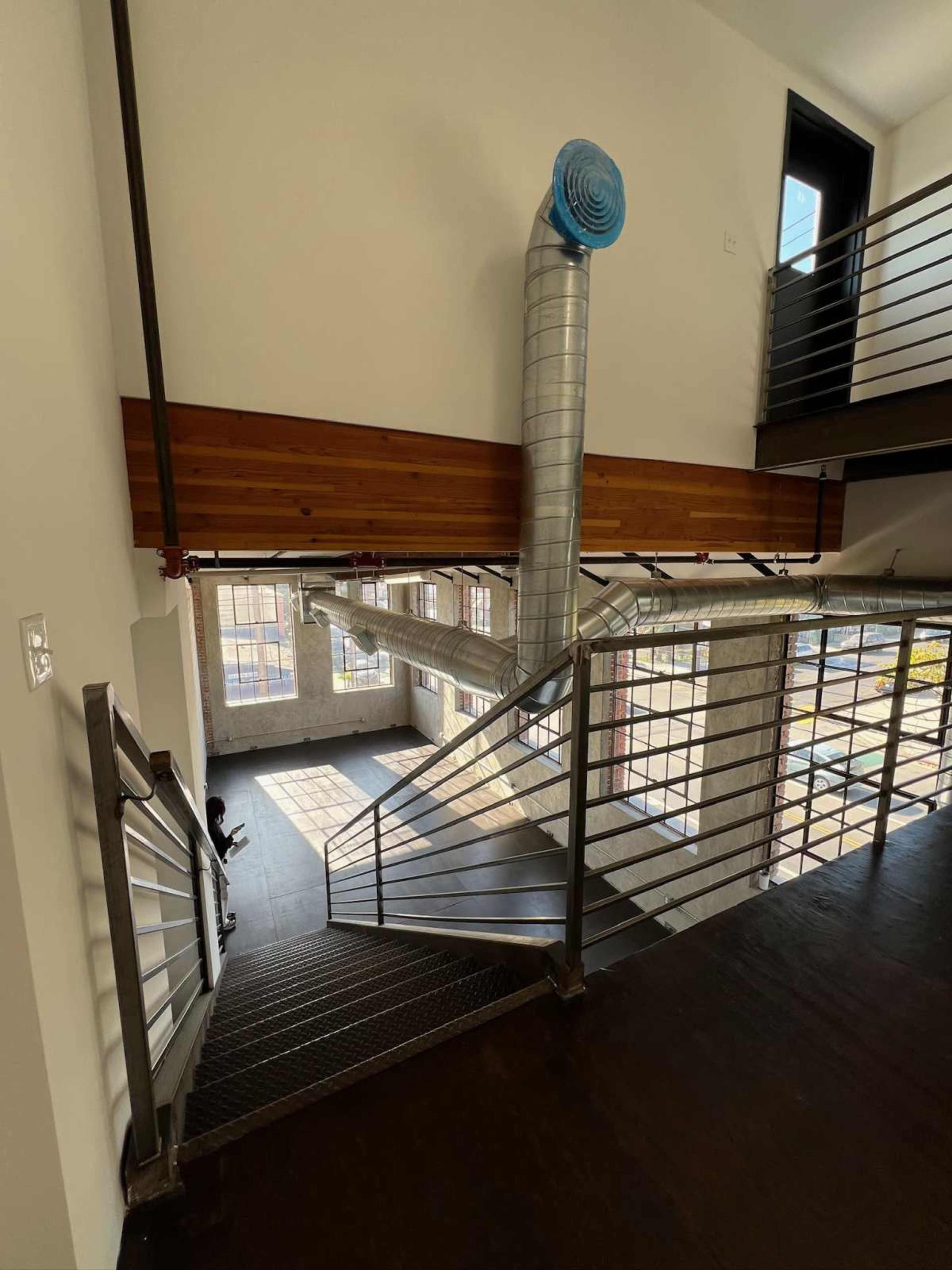 The image shows a modern interior stairway with metal railings, leading to a spacious area illuminated by natural light, featuring exposed ductwork and large windows.