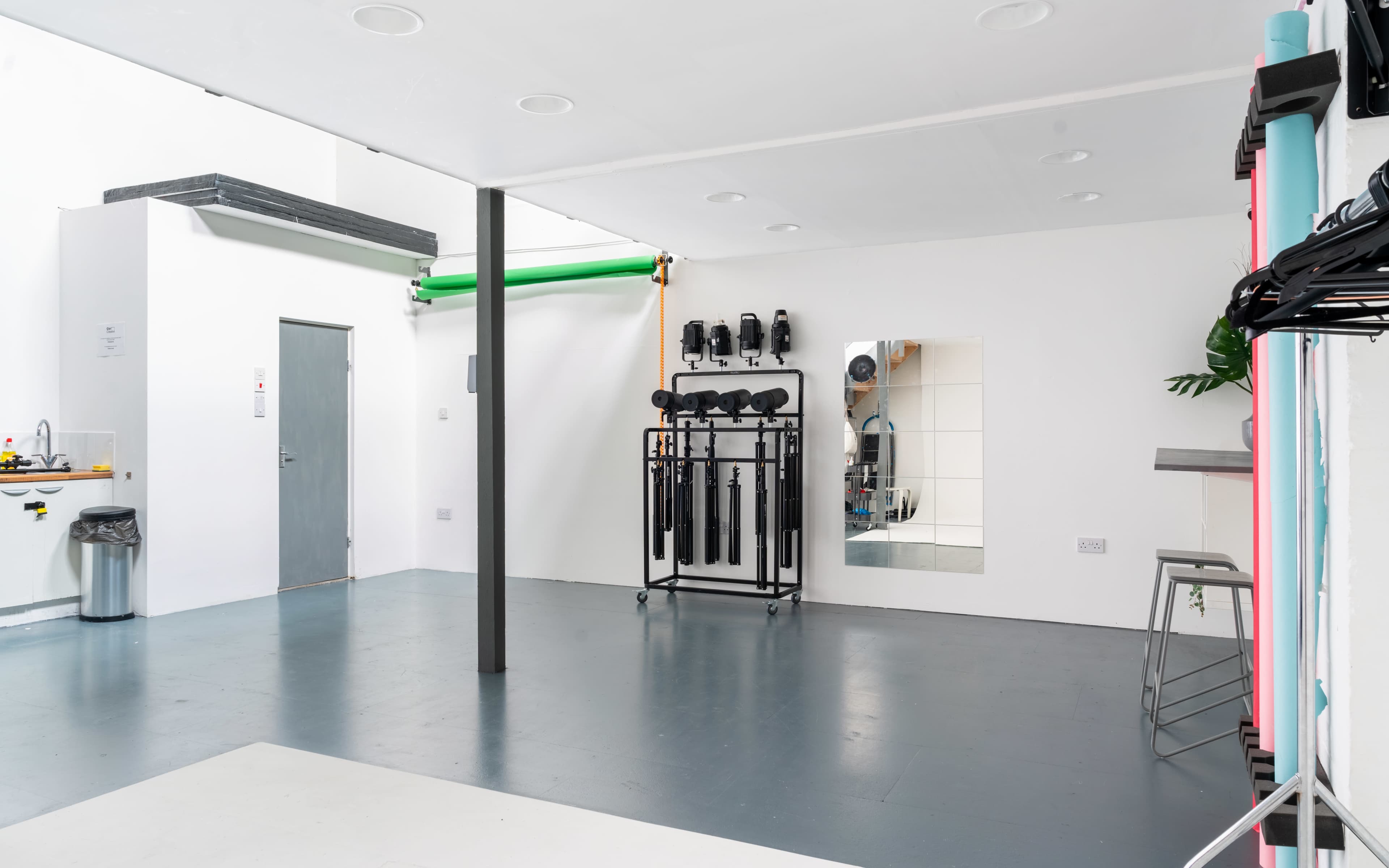 An empty studio space with a gray floor, a wall-mounted mirror, and various equipment hanging on a black mobile rack next to a door.