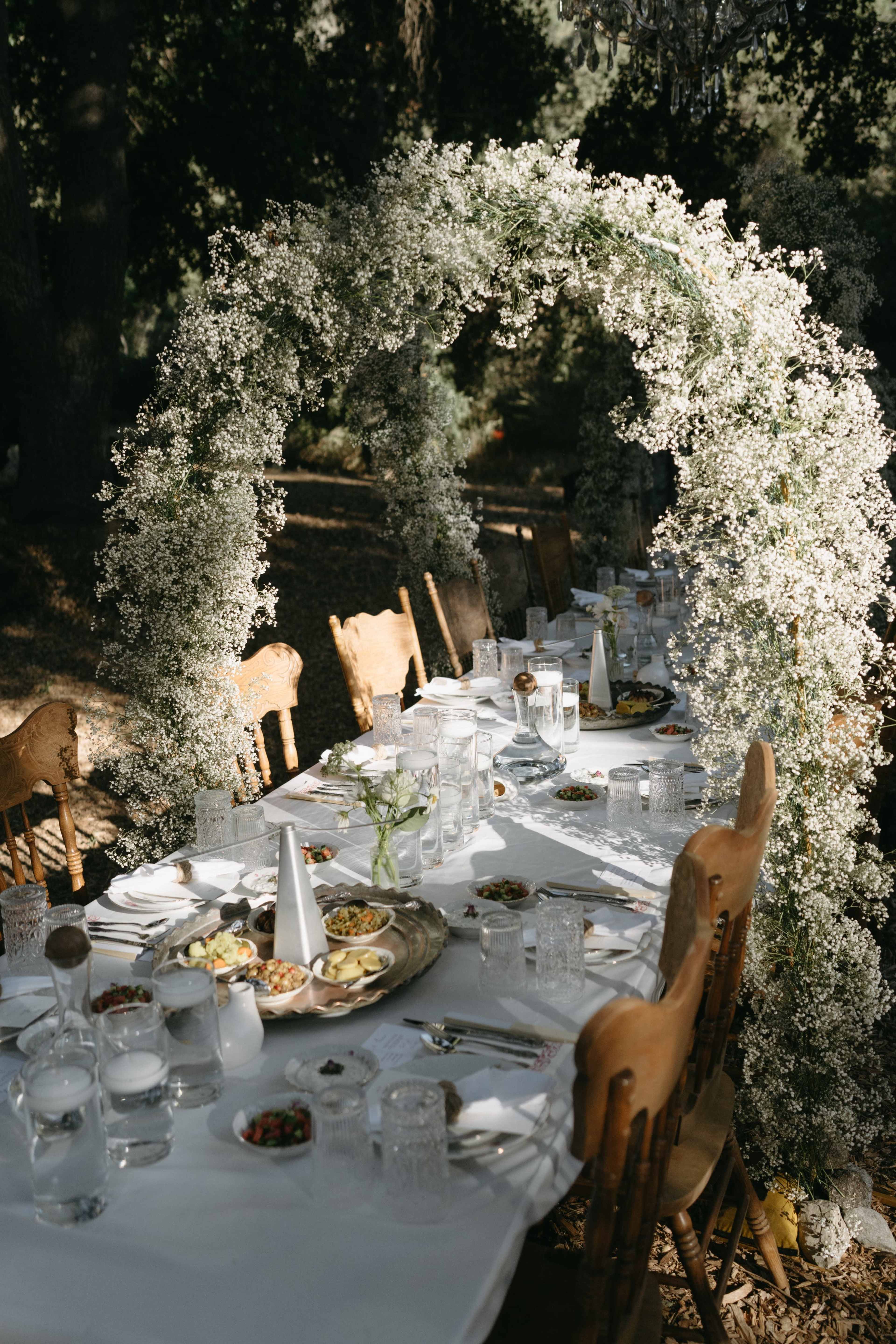 A long dining table is set outdoors under an arch made of white flowers, surrounded by chairs and adorned with various dishes and glassware.