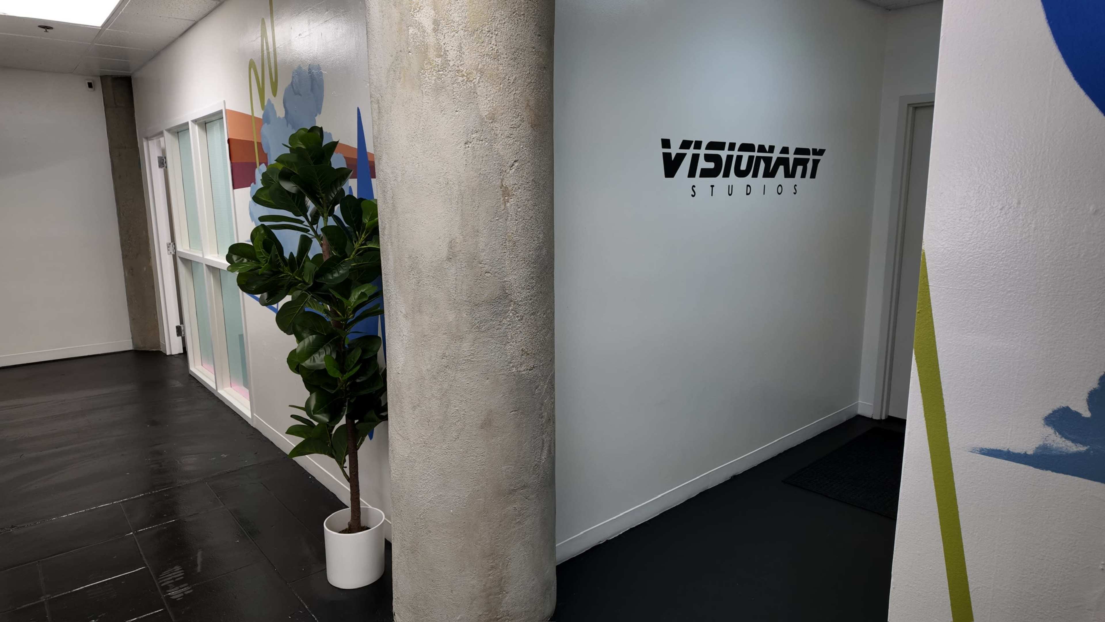 The image shows a hallway with a sign for "Visionary Studios" on a white wall, a potted plant beside a concrete pillar, and colorful wall graphics in the background.
