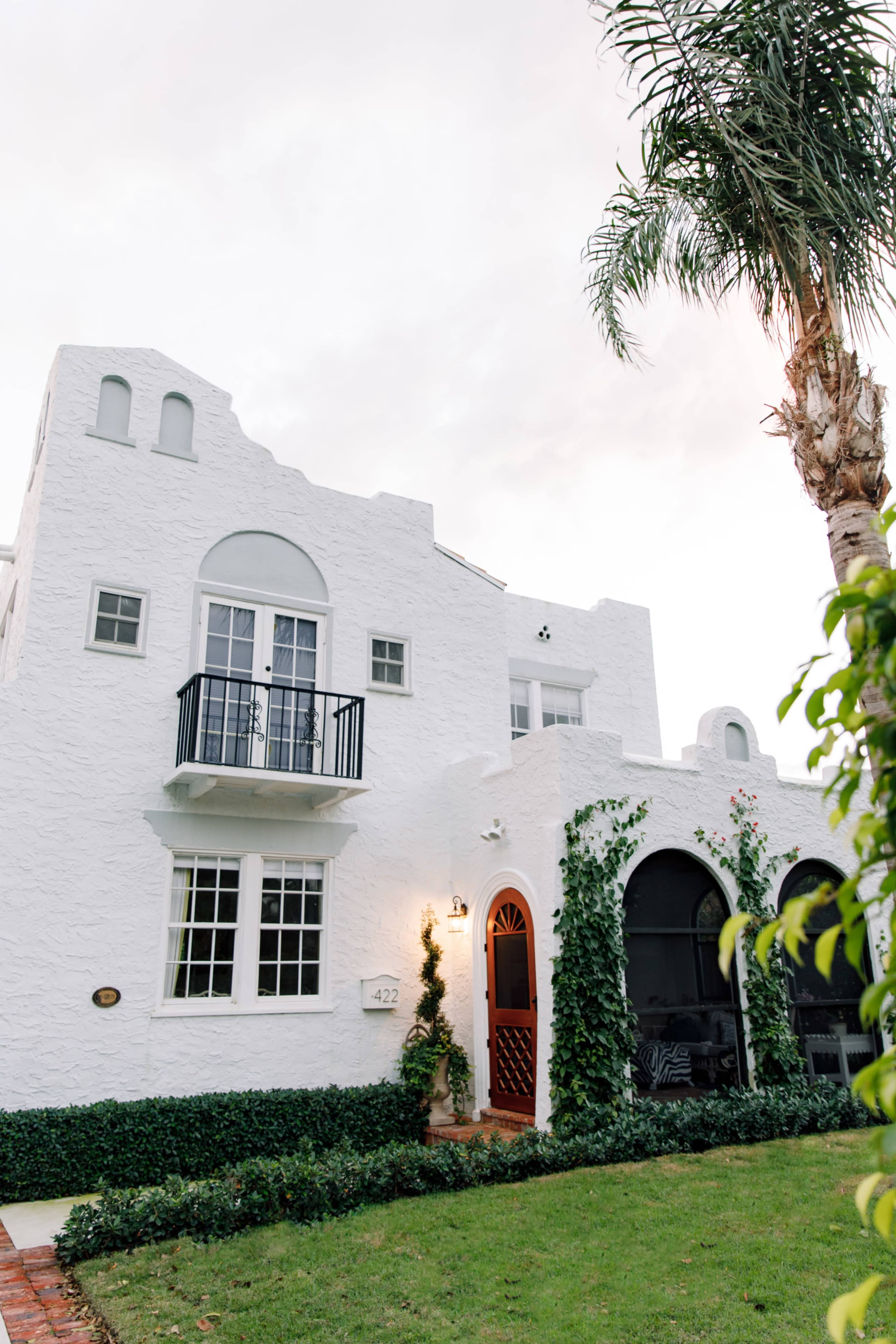 The image shows a white, textured house with a black balcony and arched entrances, surrounded by lush greenery and a palm tree.