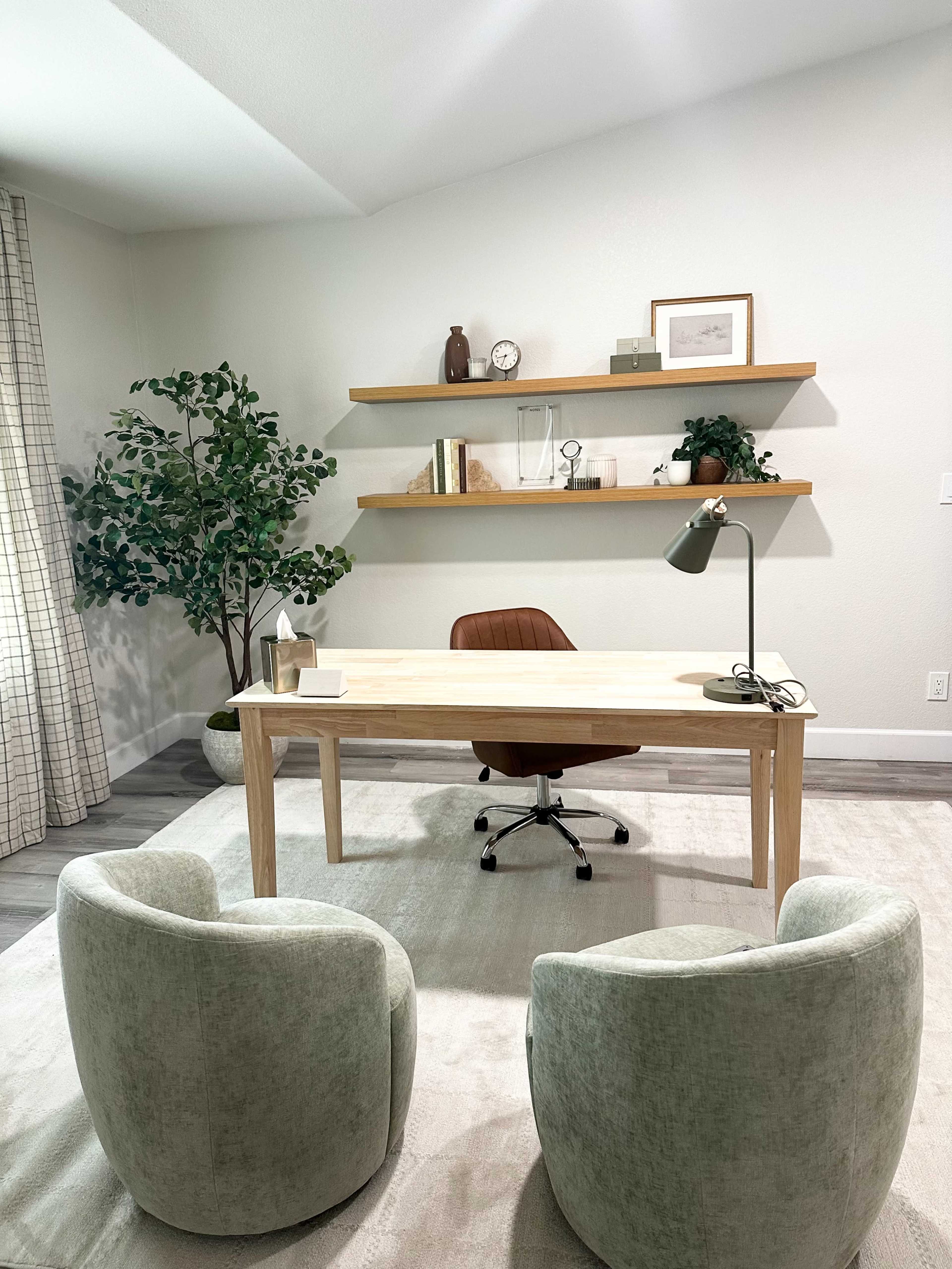 A modern office space features a wooden desk with a chair, two upholstered chairs, and floating shelves holding decorative items and books.