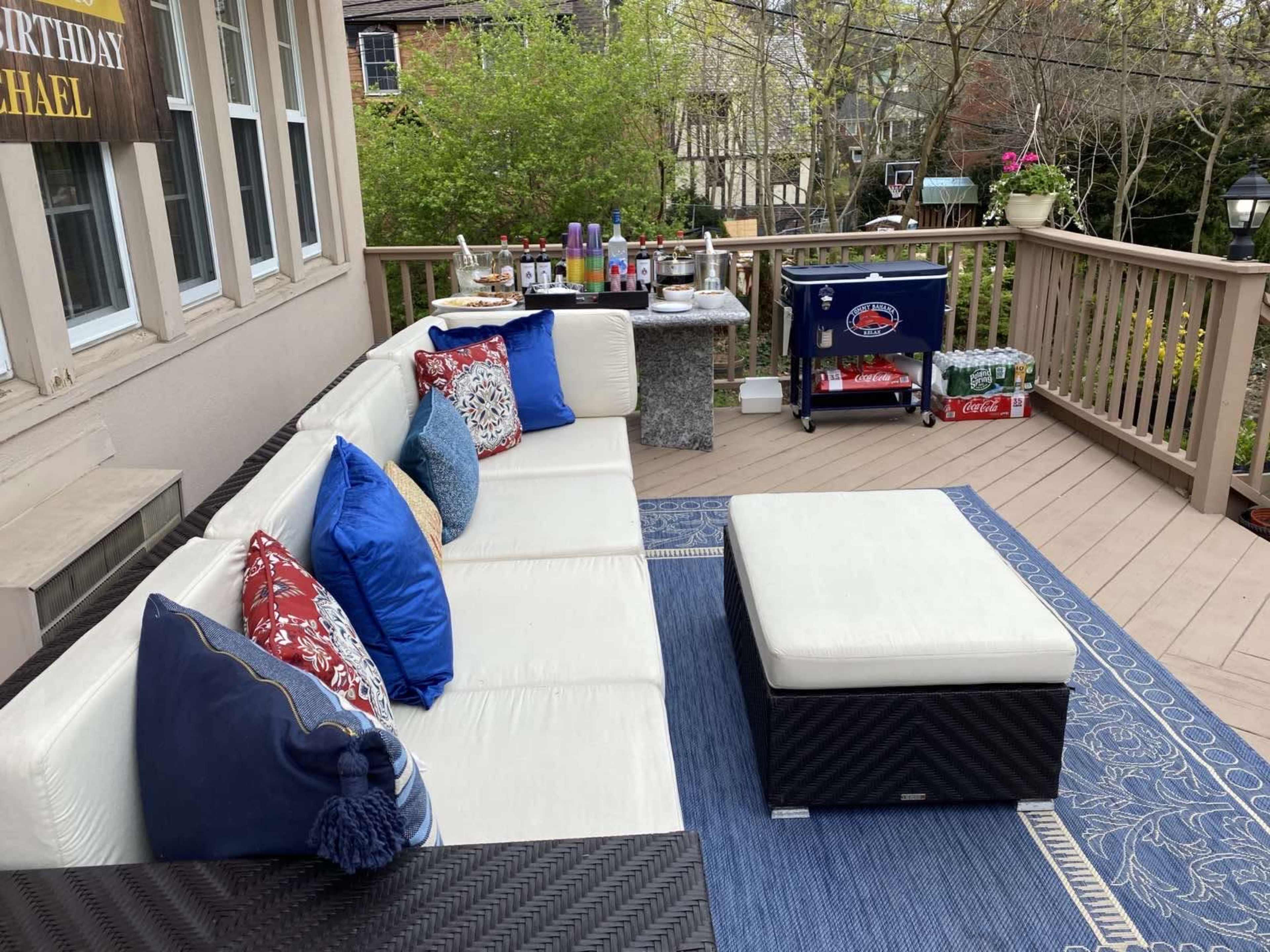 A spacious outdoor deck features a white sectional sofa with colorful pillows, a coffee table, and a bar area stocked with drinks and a cooler in the background.