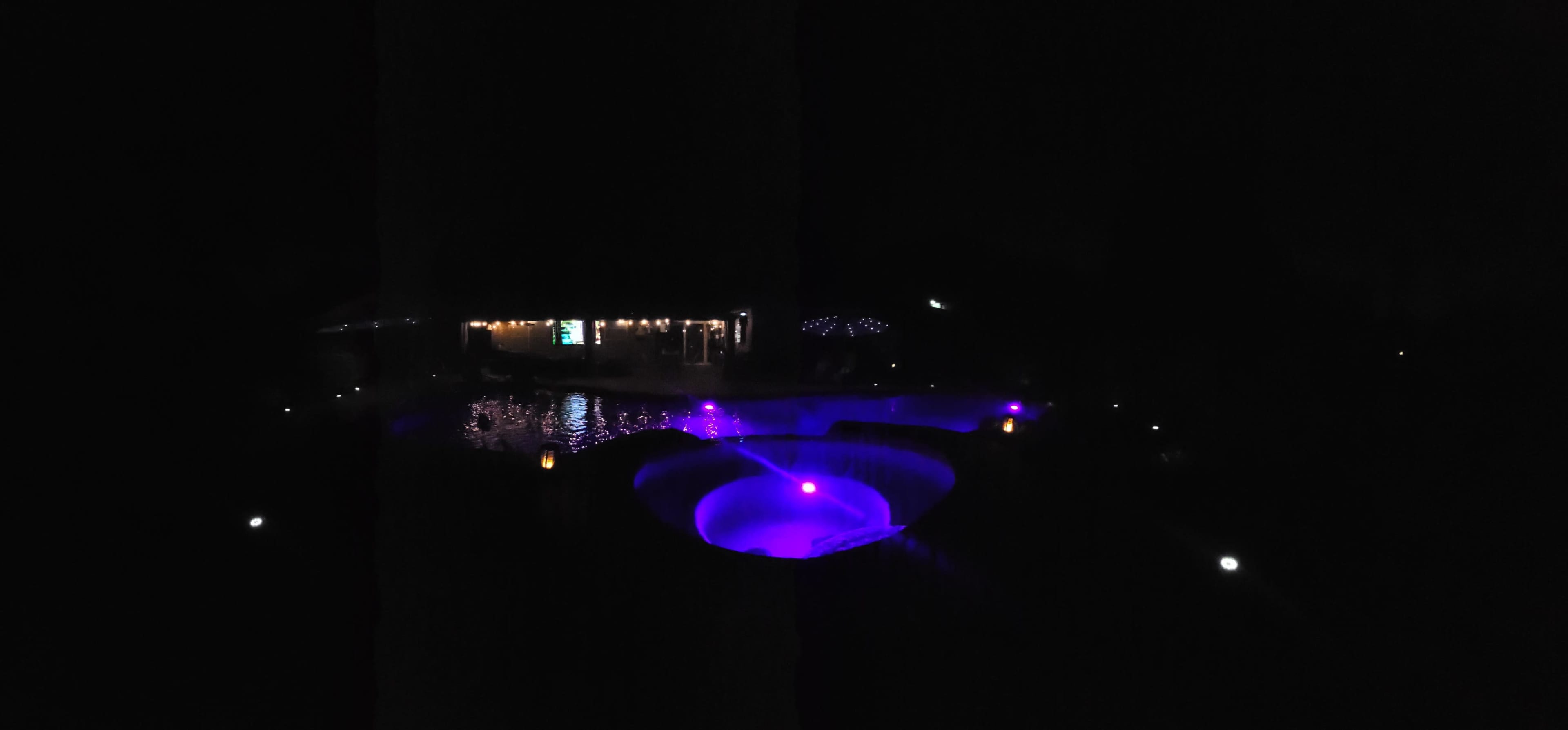 A pool with purple lighting is surrounded by trees and a building at night.