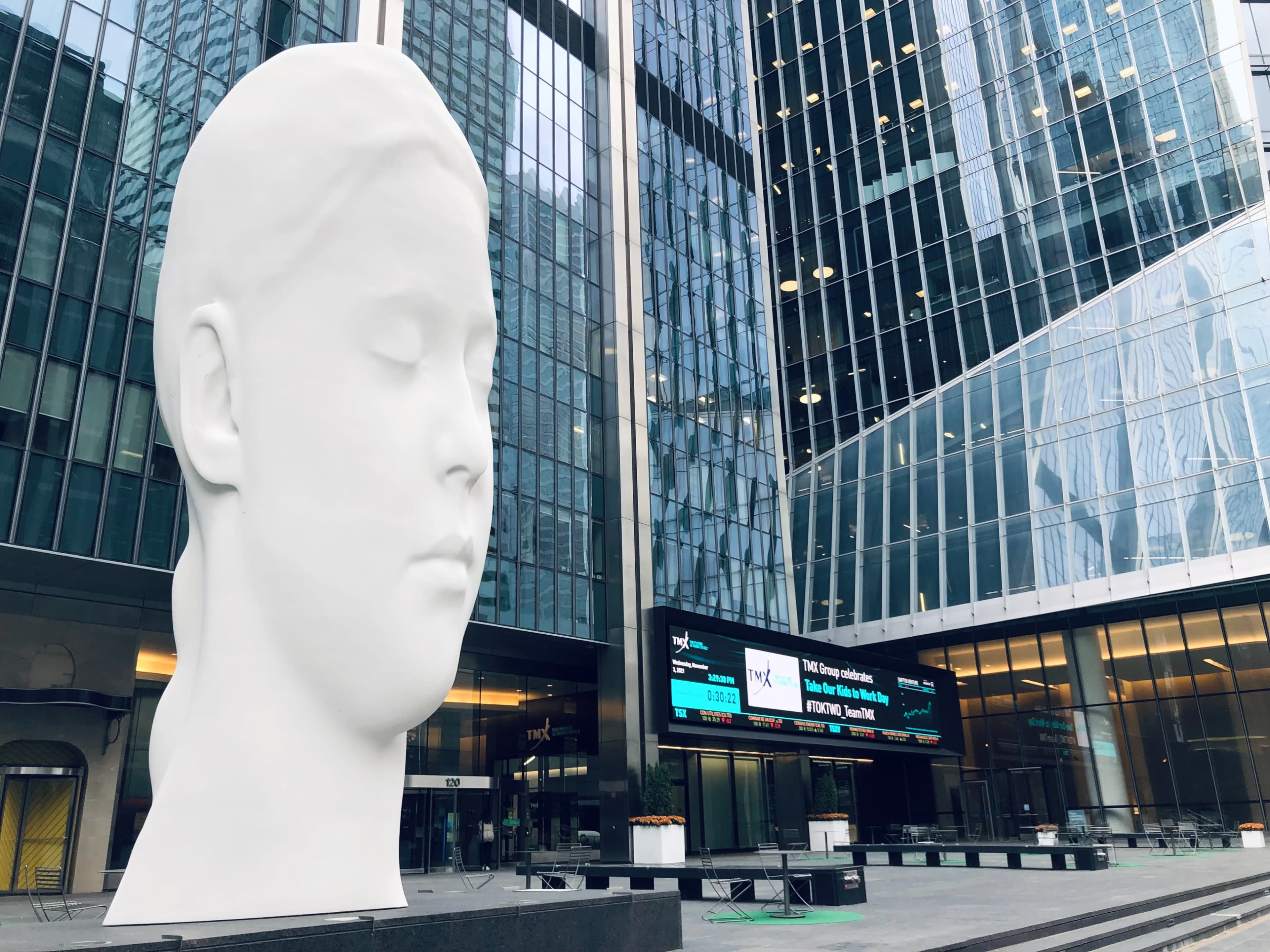 A large white sculpture of a female face sits in an urban plaza surrounded by glass buildings and a digital billboard.
