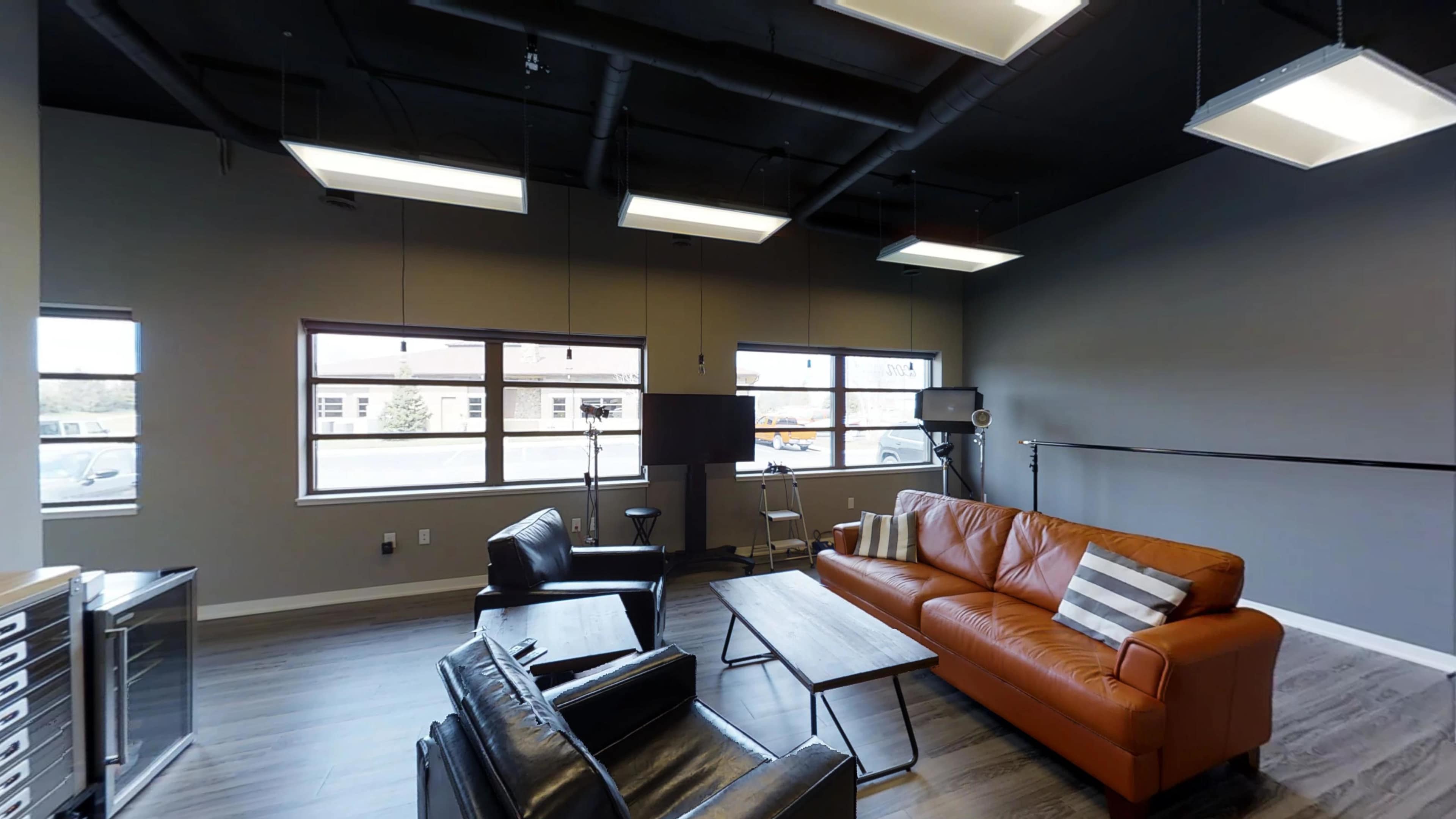 A modern, well-lit office lounge area featuring a brown leather sofa, a wooden coffee table, and several windows.