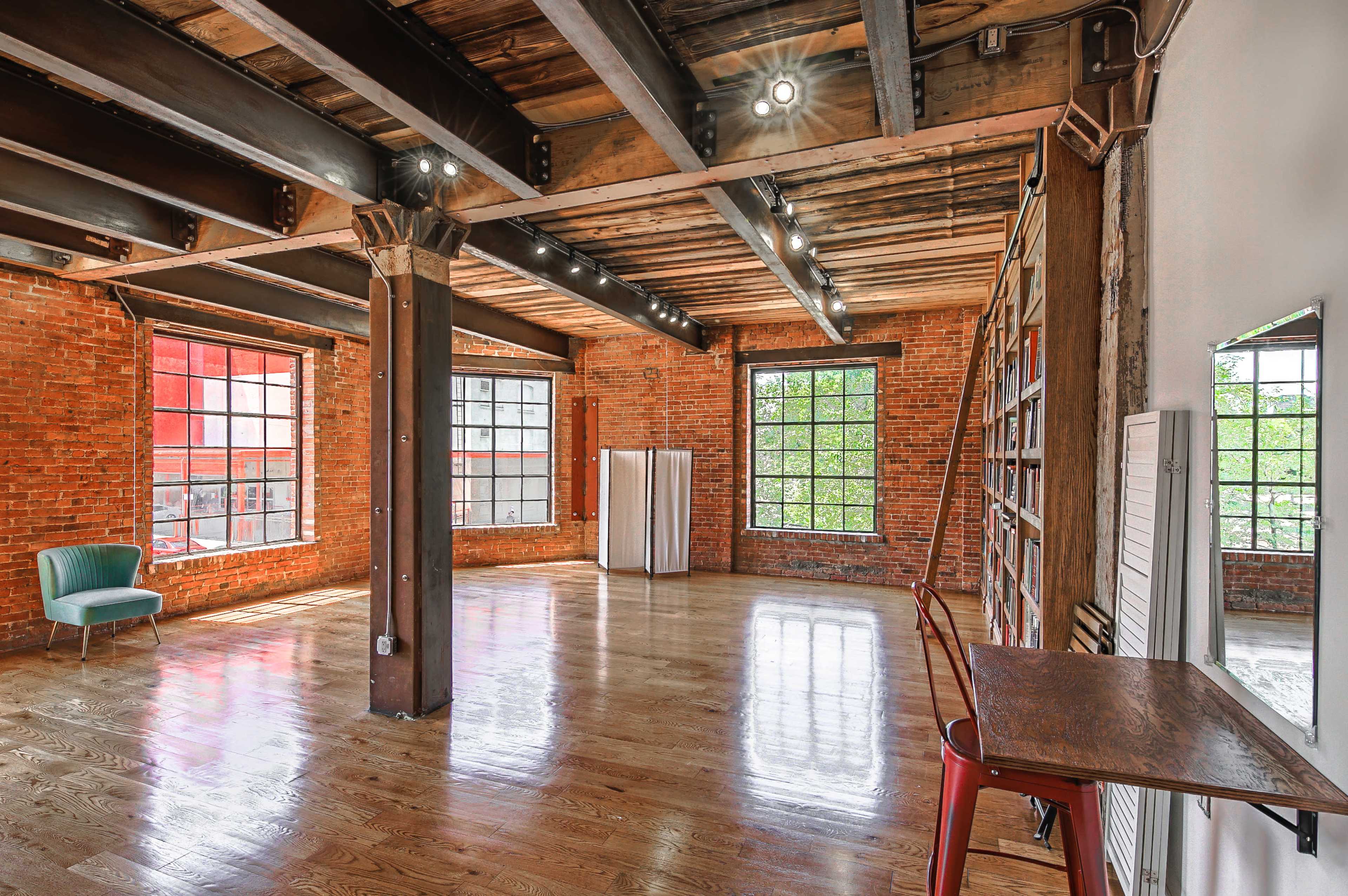 The image shows a spacious room with brick walls, wooden beams, large windows, and polished wooden floors, featuring a green chair and a table against a wall.