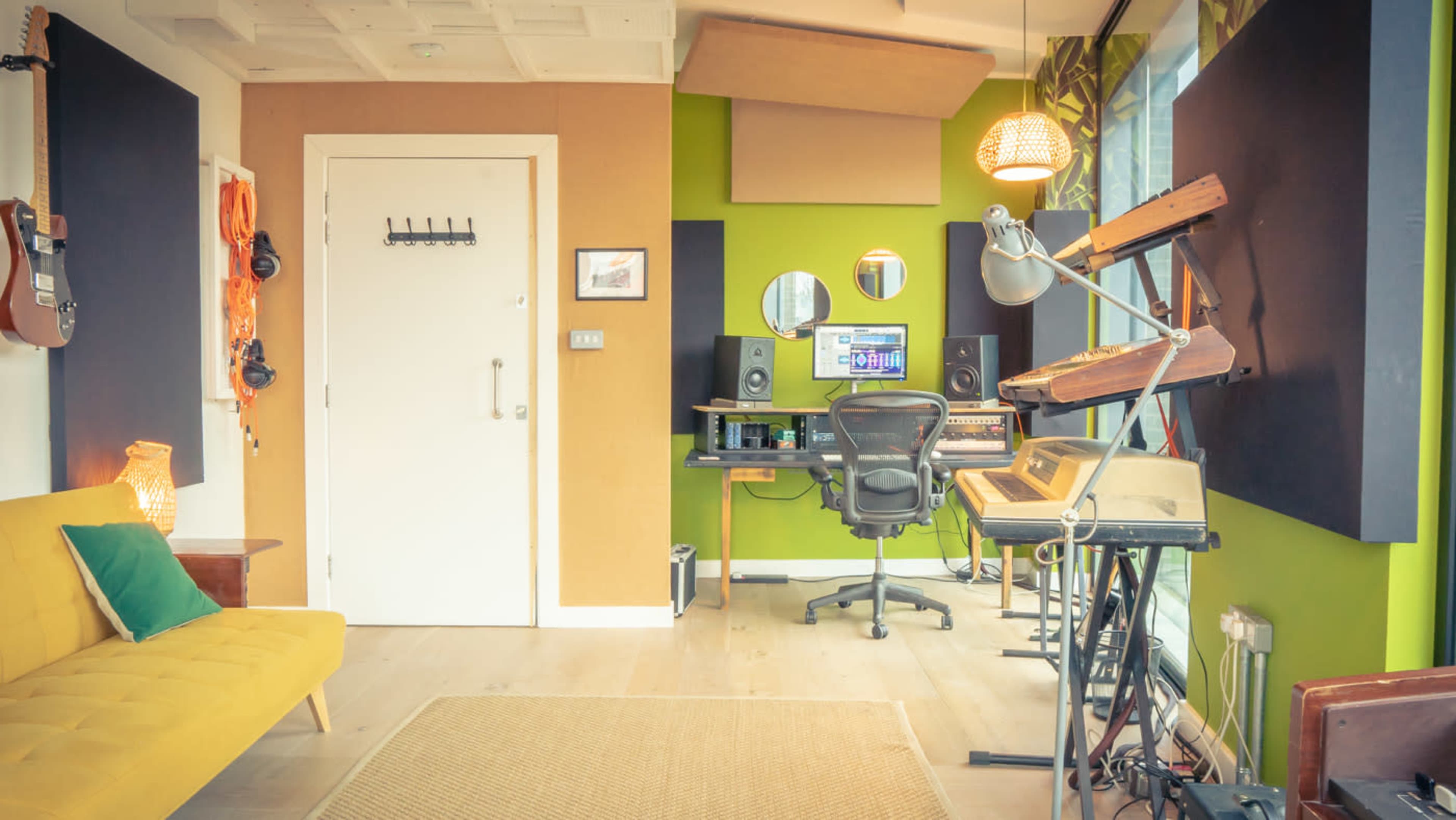 A small music studio features a workstation with speakers and a computer, surrounded by acoustic panels and musical instruments.