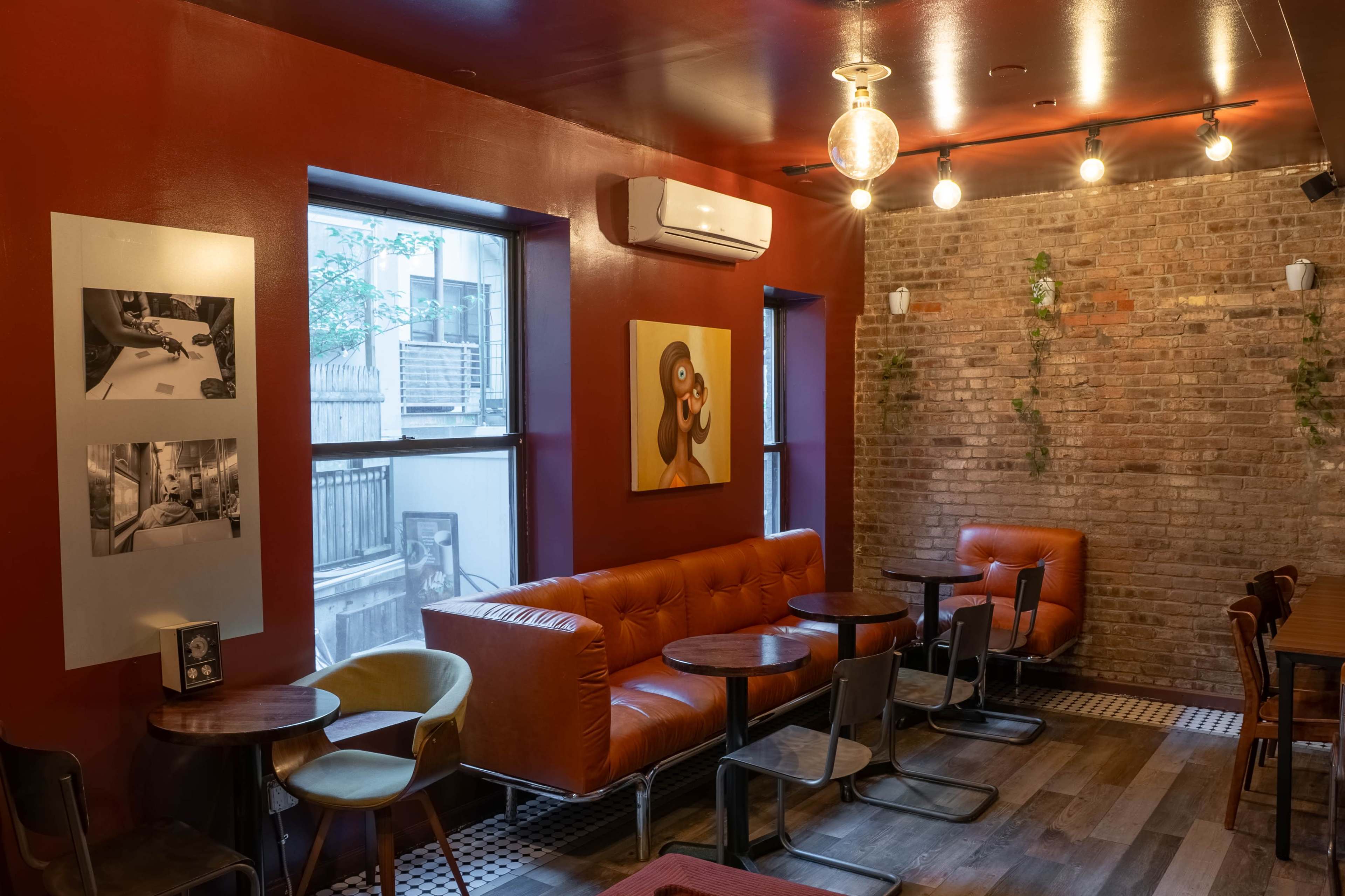 The interior of a cozy cafe features orange walls, a brick accent wall, leather seating, small tables, and decorative artwork.
