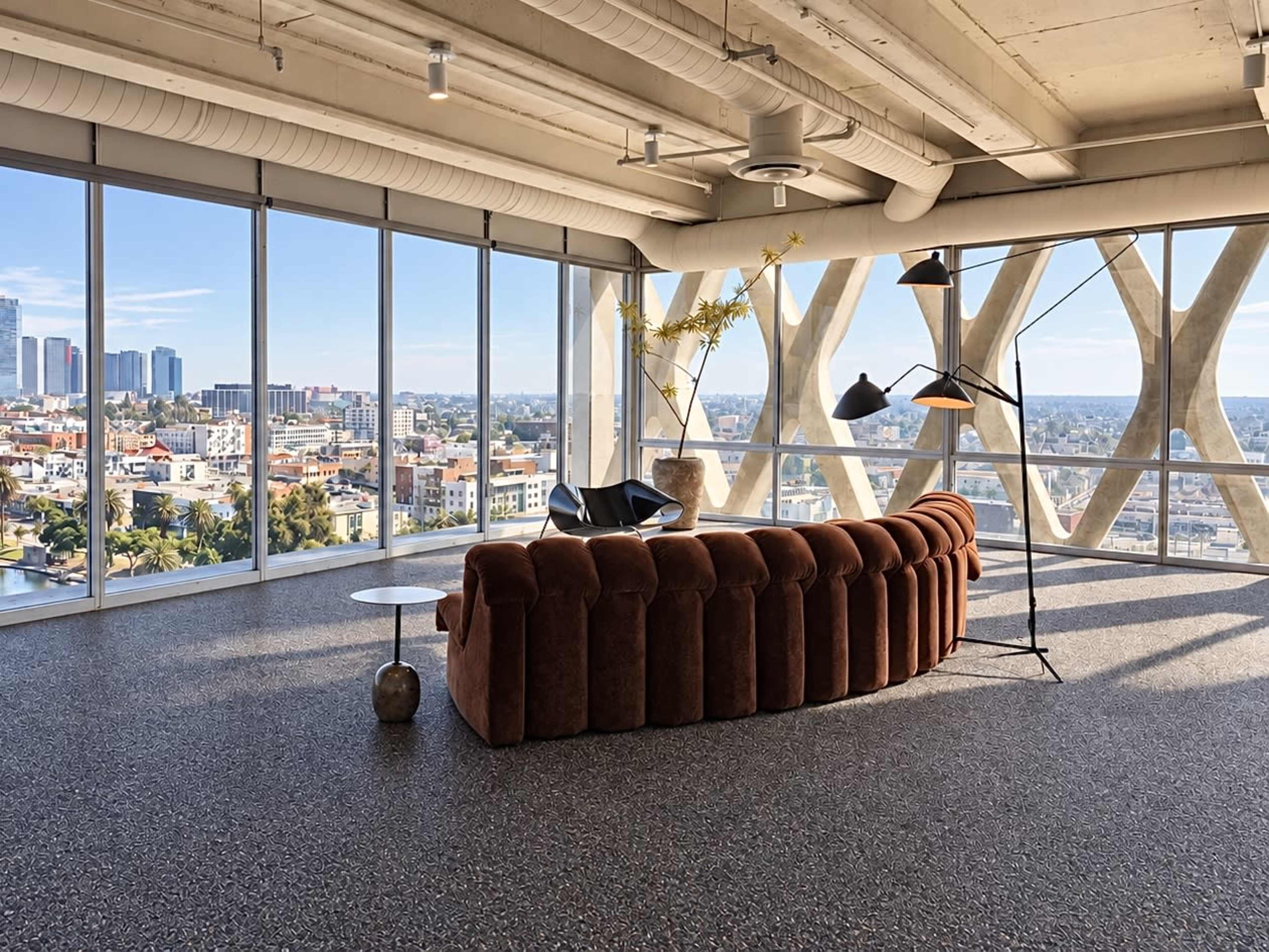 The image shows a modern, spacious interior with large windows offering a view of a city skyline, featuring a curved brown sofa, a side table, and a floor lamp.