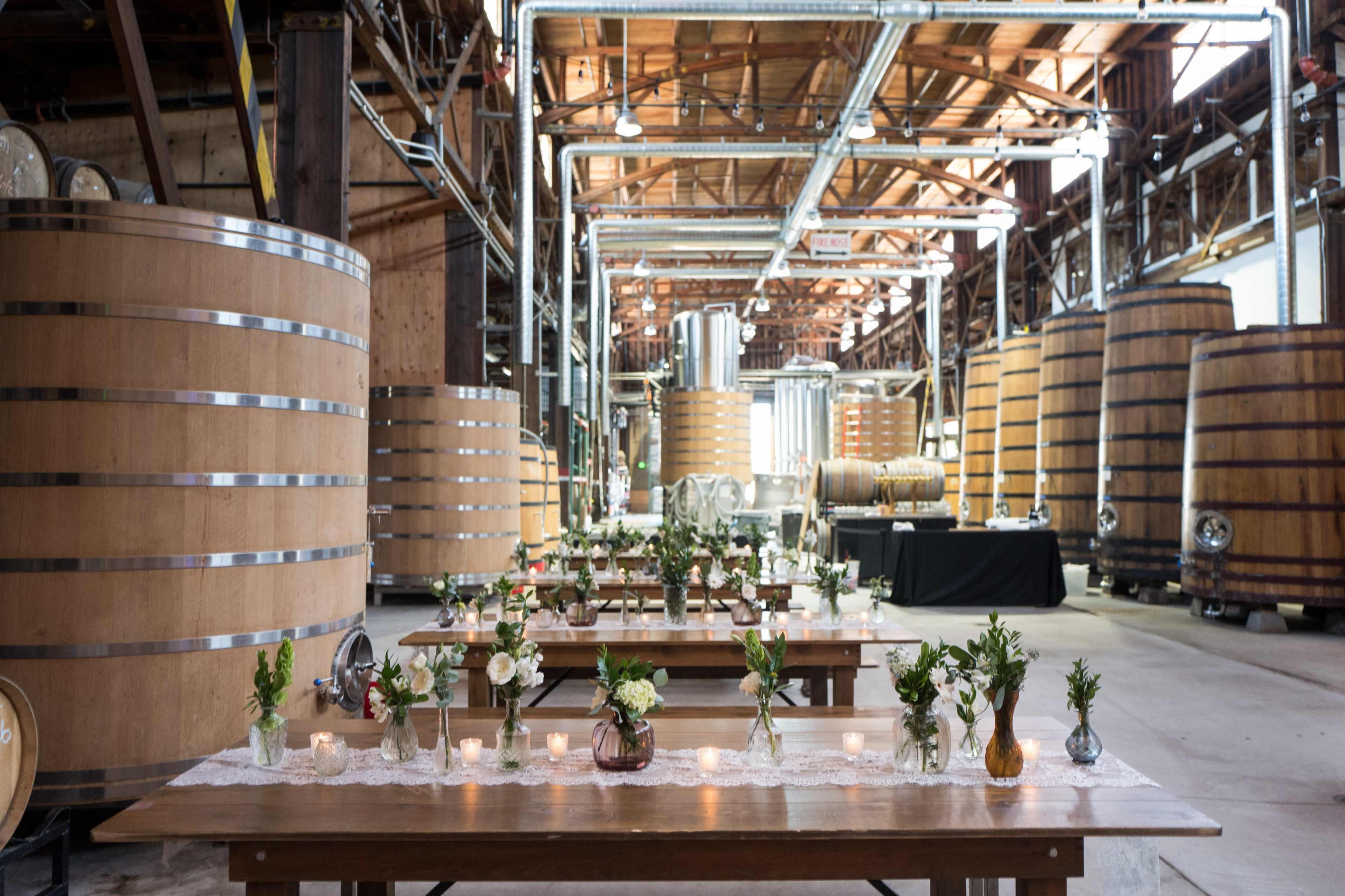 The image shows a spacious winery interior with large wooden fermentation tanks and elegantly arranged tables set with flowers and candles.