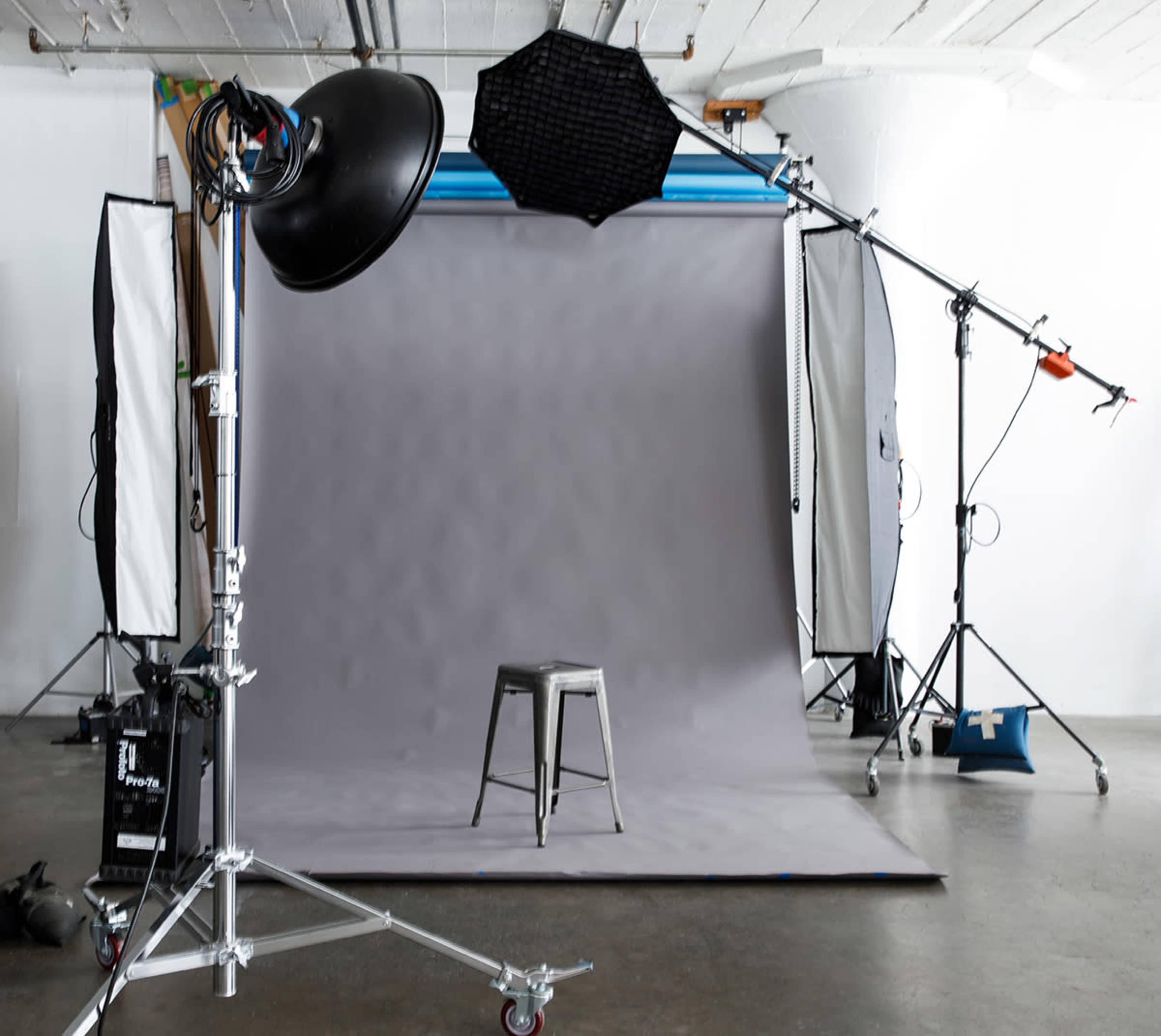 The image shows a photography studio setup with a gray backdrop, a stool in the center, and various lighting equipment arranged around it.