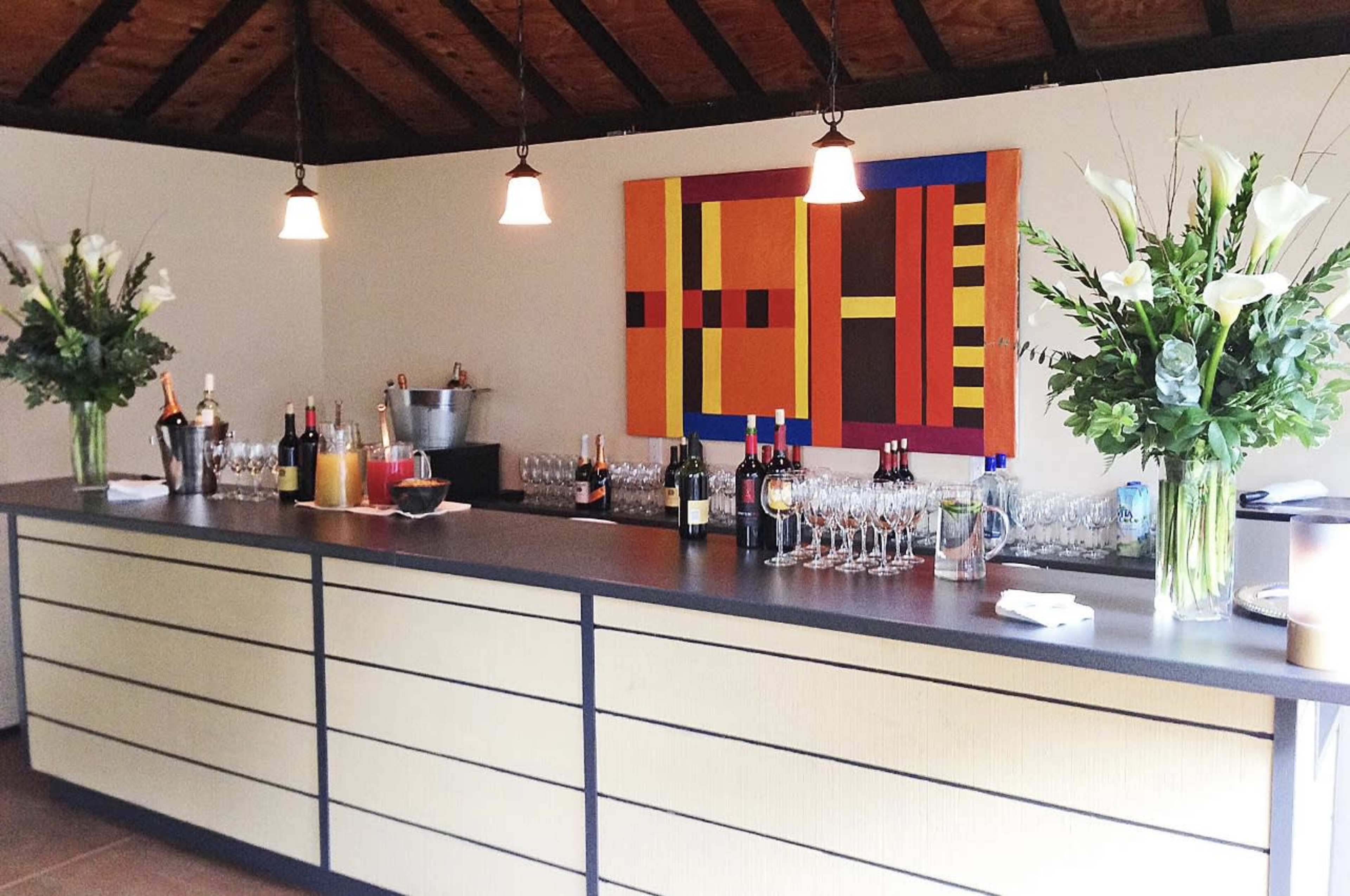 A bar area features a long counter with various bottles and glasses arranged alongside large floral arrangements and an abstract painting on the wall.