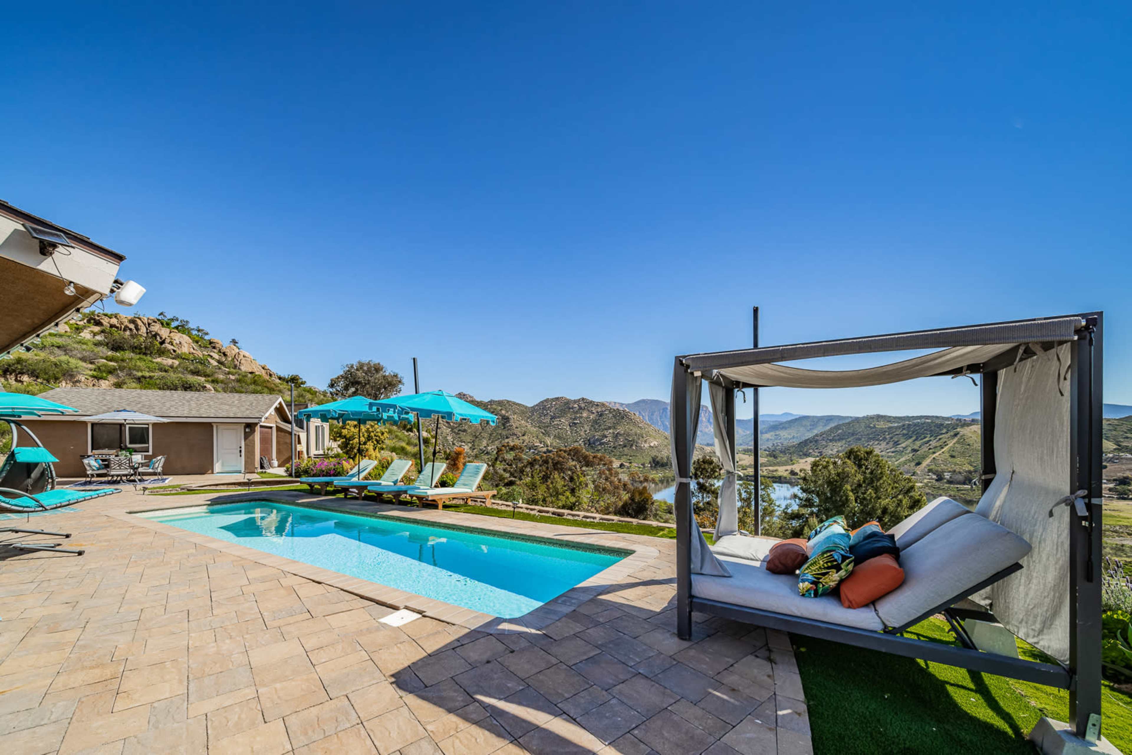 A modern outdoor pool area features a lounging bed, sun loungers, and umbrellas, set against a backdrop of rolling hills and a lake.