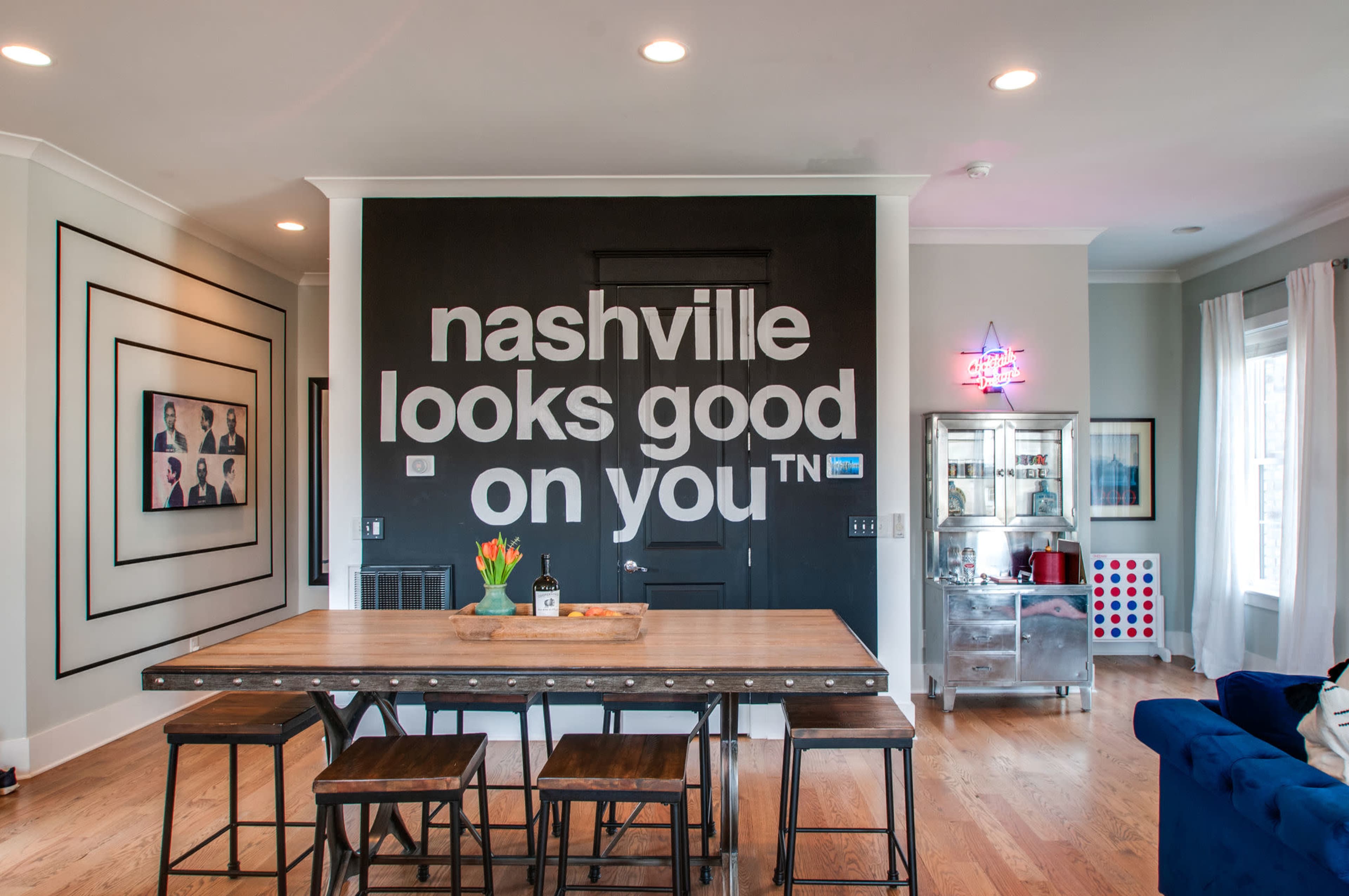 A modern dining area with a large wooden table and a prominent black wall featuring the phrase "nashville looks good on you" in white letters.