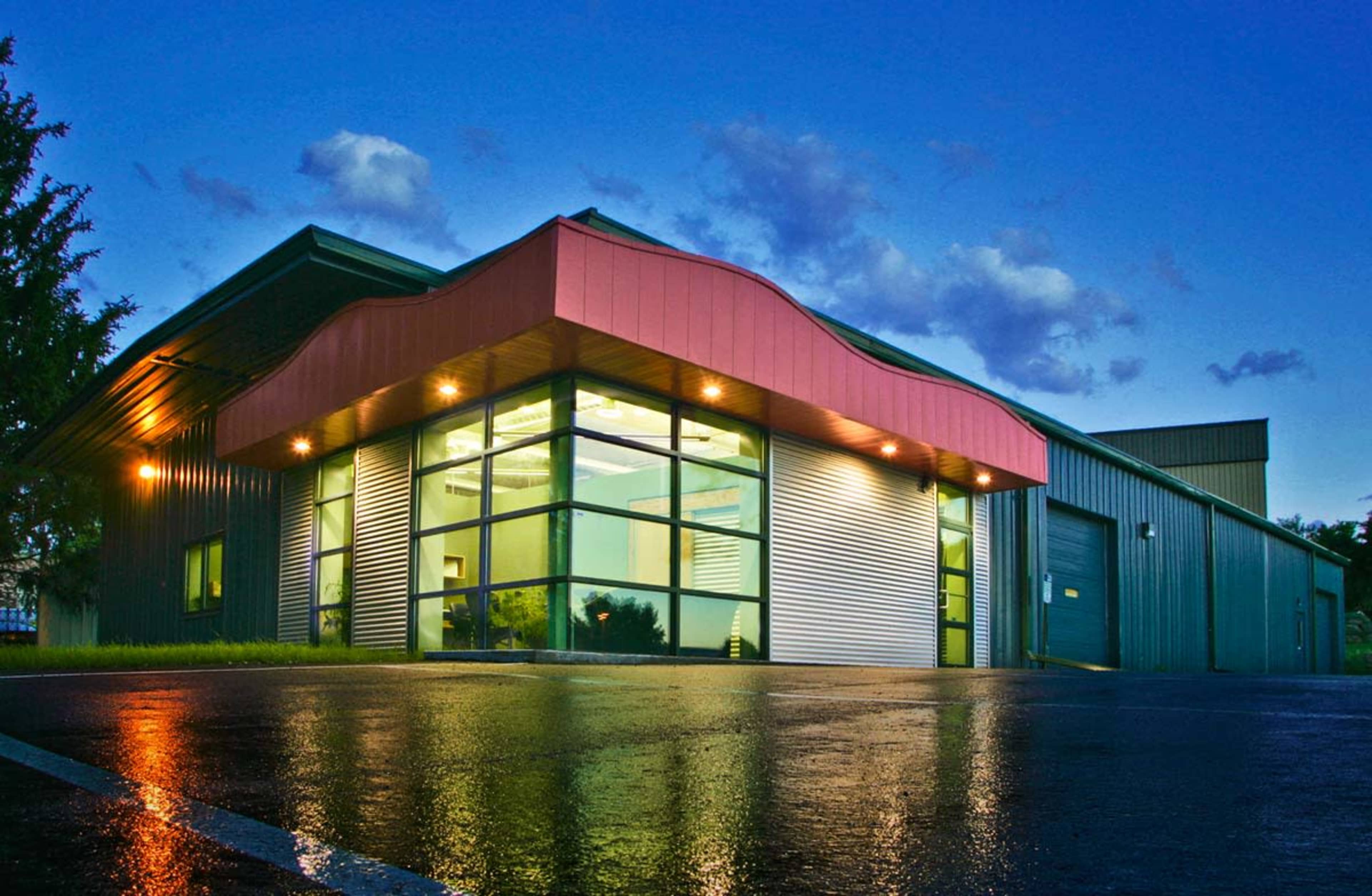 A modern commercial building with a curved red roof and large glass windows reflects light on a wet pavement during twilight.