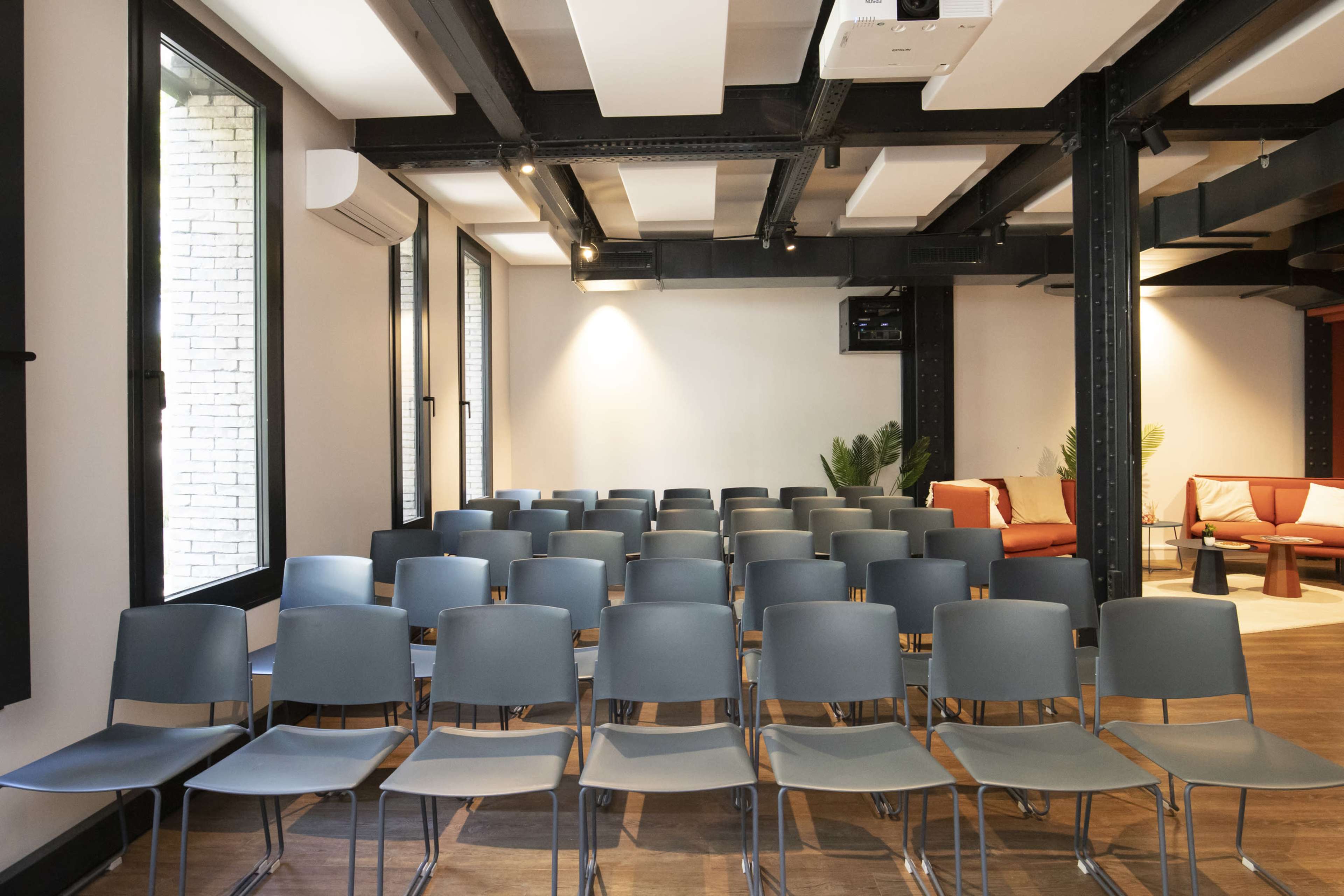 A row of empty blue chairs is arranged in neat rows in a modern room with large windows and a seating area in the background.