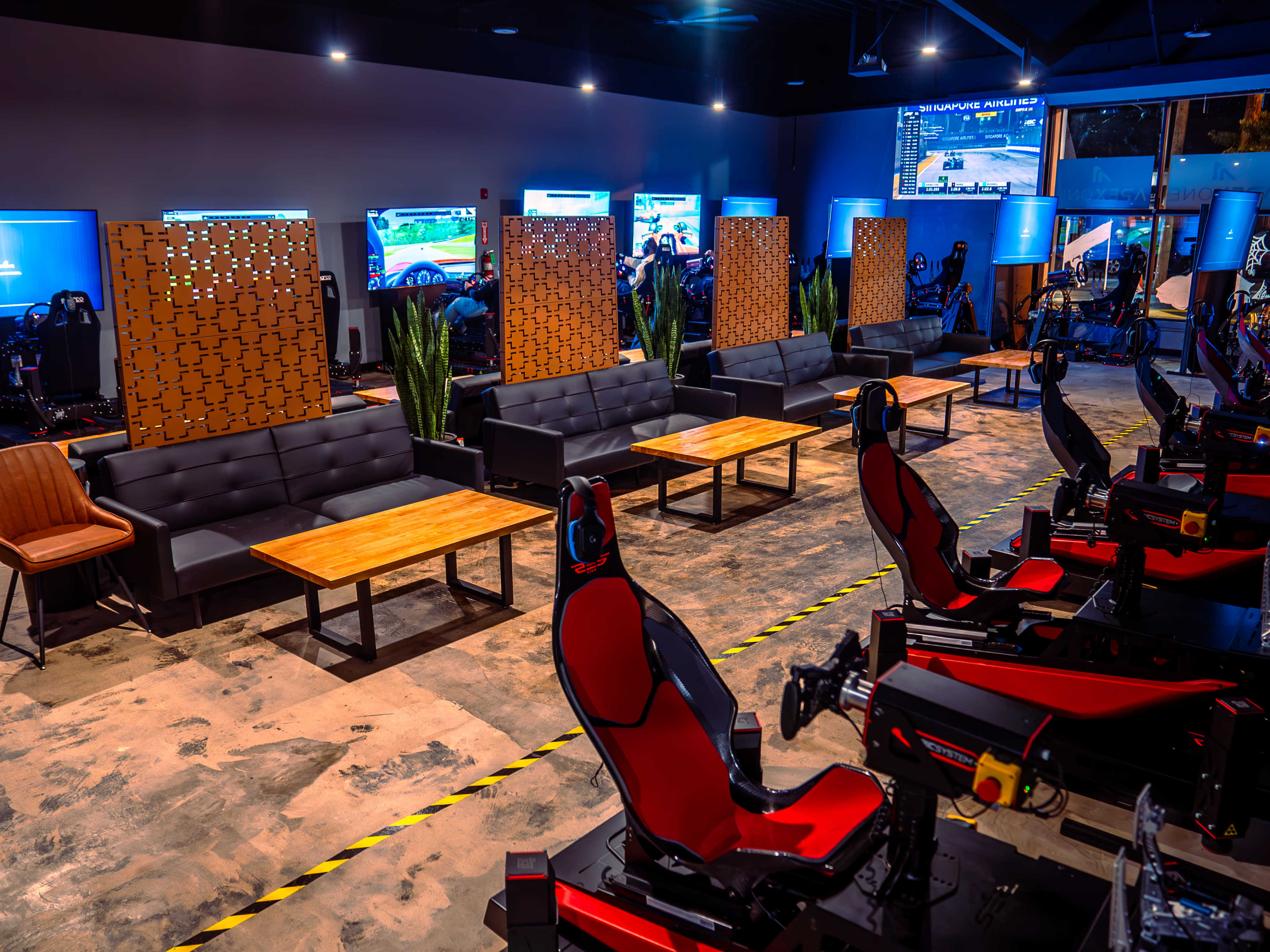 The image depicts a modern indoor racing simulation center with multiple racing seats facing screens, surrounded by lounge seating and decorative partitions.