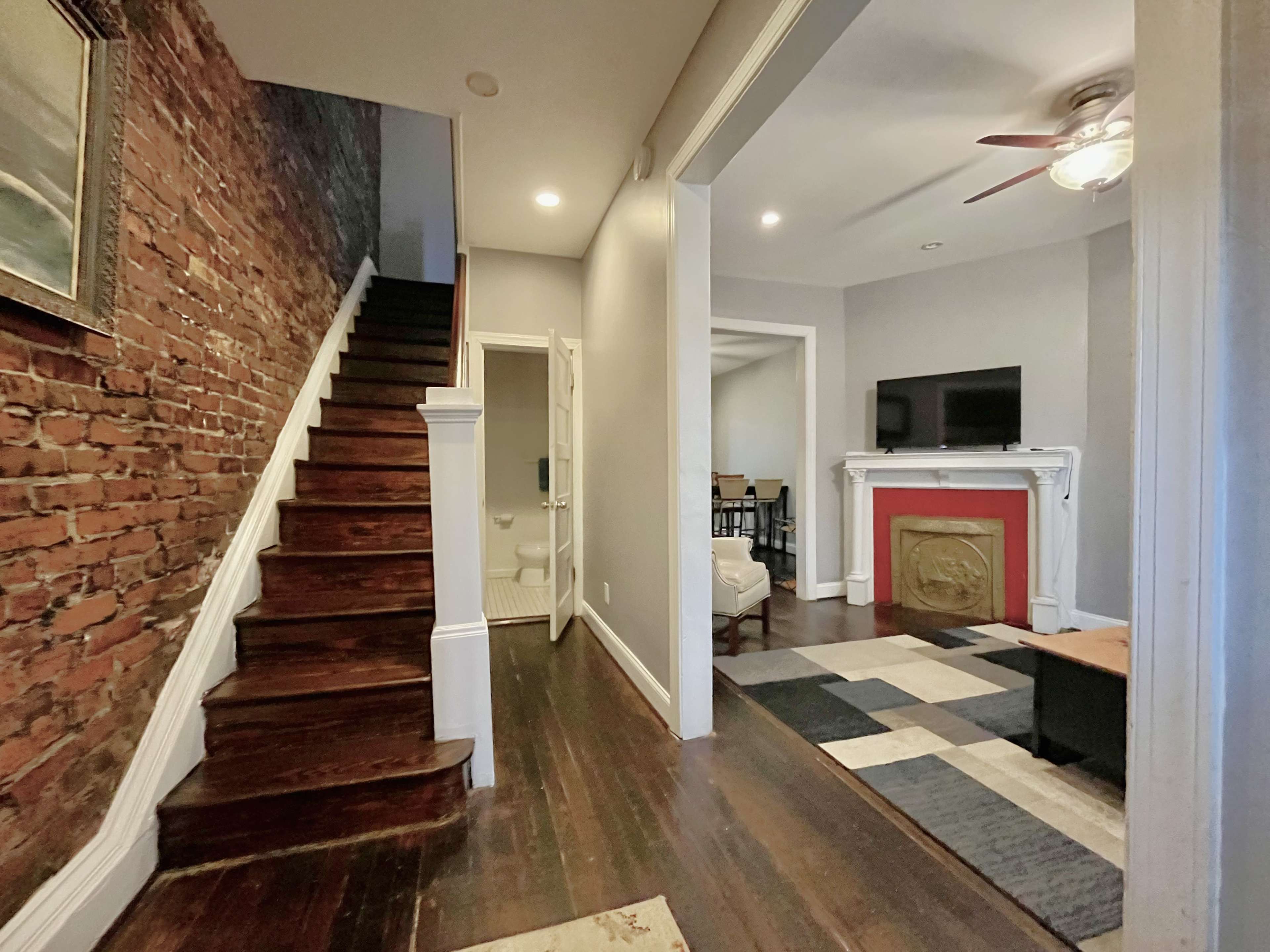 A hallway with exposed brick walls, a staircase leading upward, and a living area featuring a television on a mantel with a red surround.