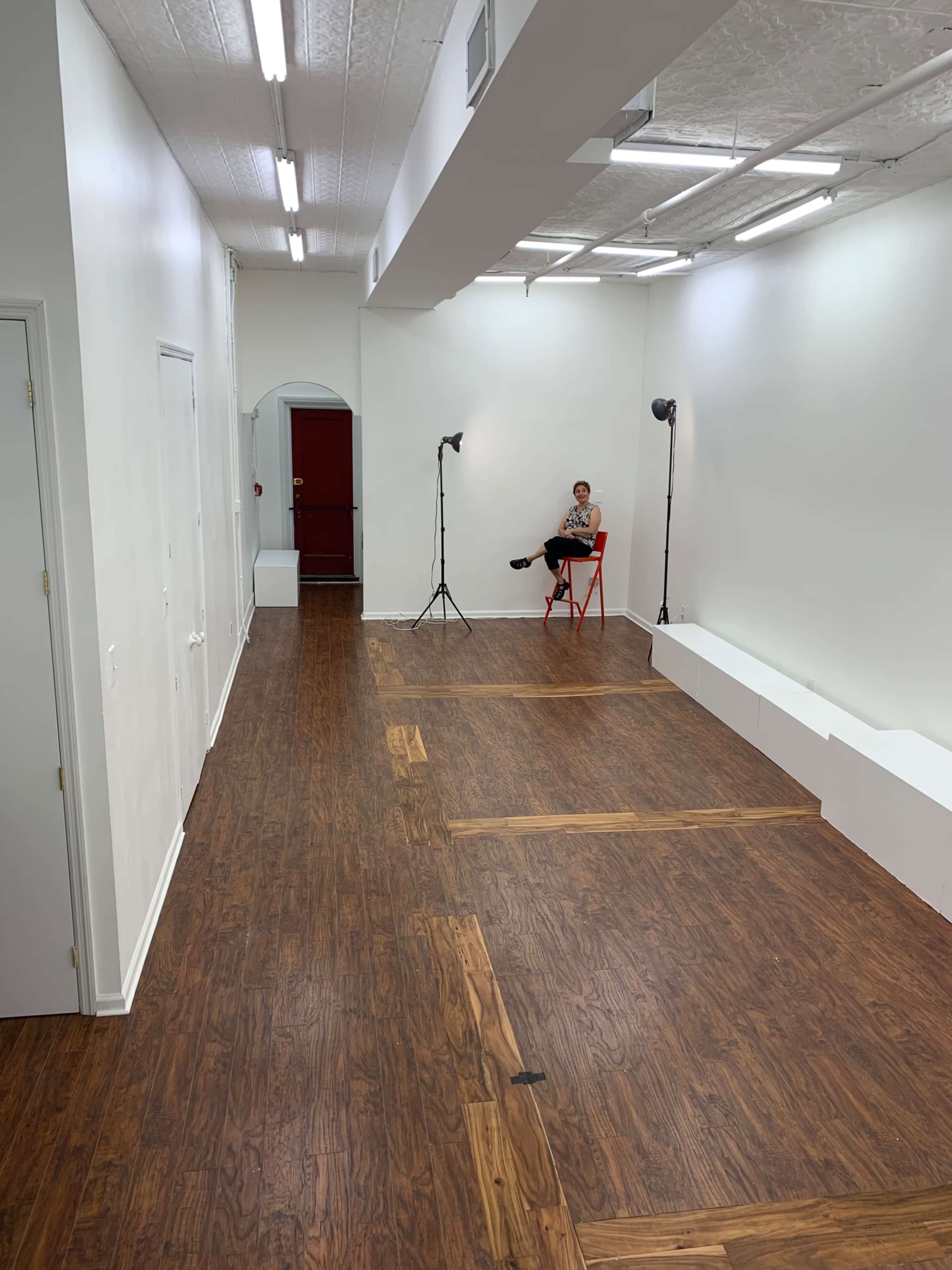 A spacious, brightly lit room with wooden flooring features a single person seated on a red chair surrounded by studio lighting equipment.