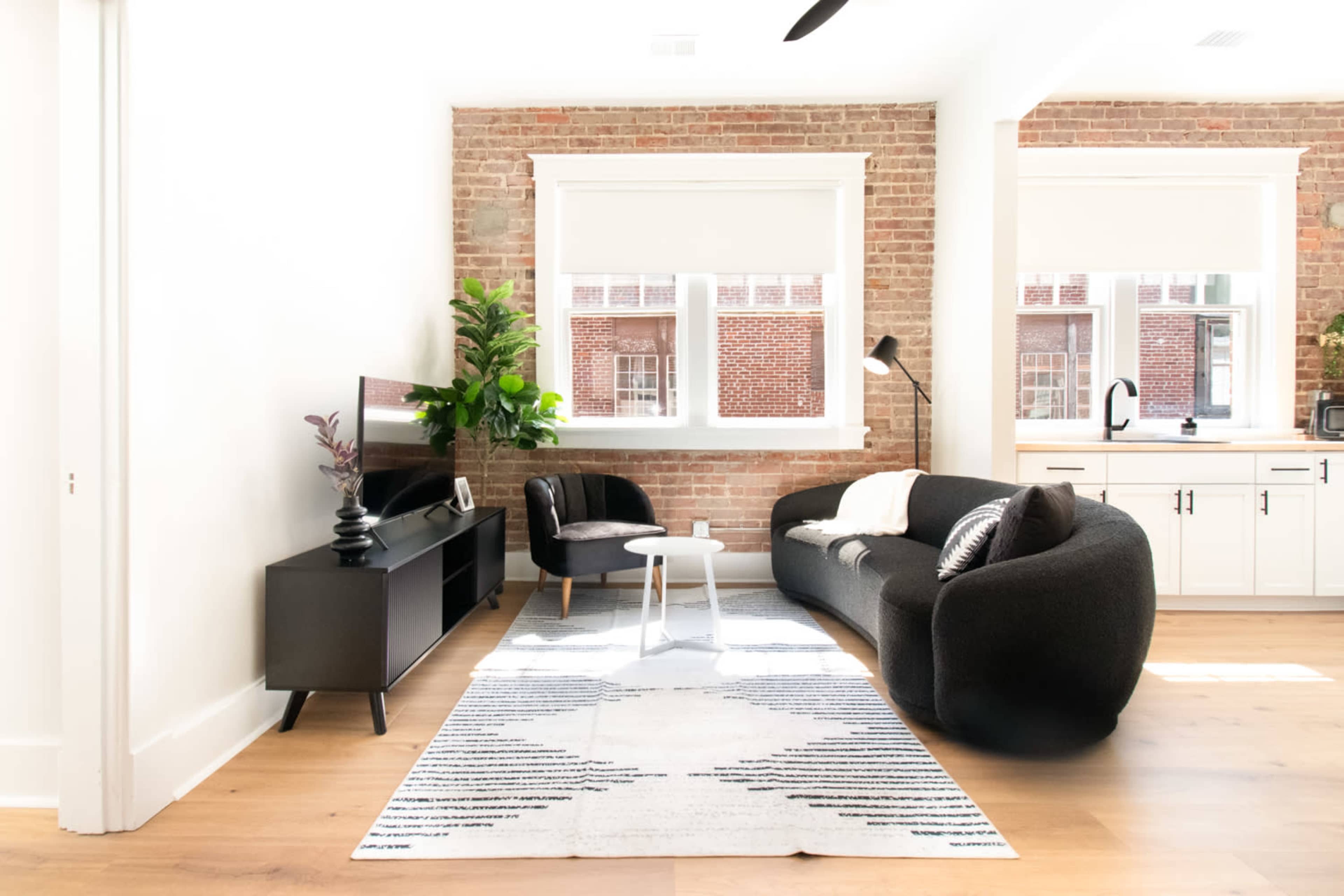 A modern living room features a curved black sofa, a small round table, and a potted plant, against a backdrop of exposed brick walls and large windows.
