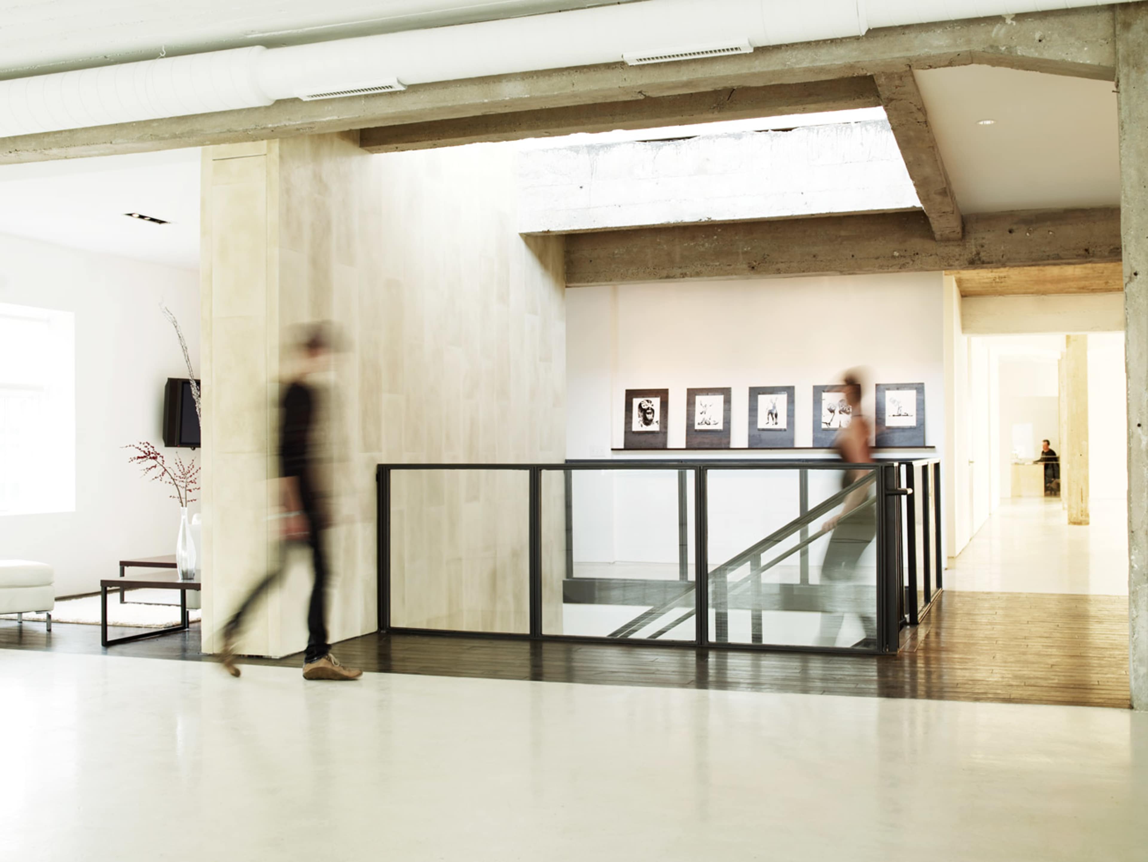 The image shows a modern interior space with a staircase, large windows, and framed artwork on the walls, while two people walk past each other in a blurred motion.