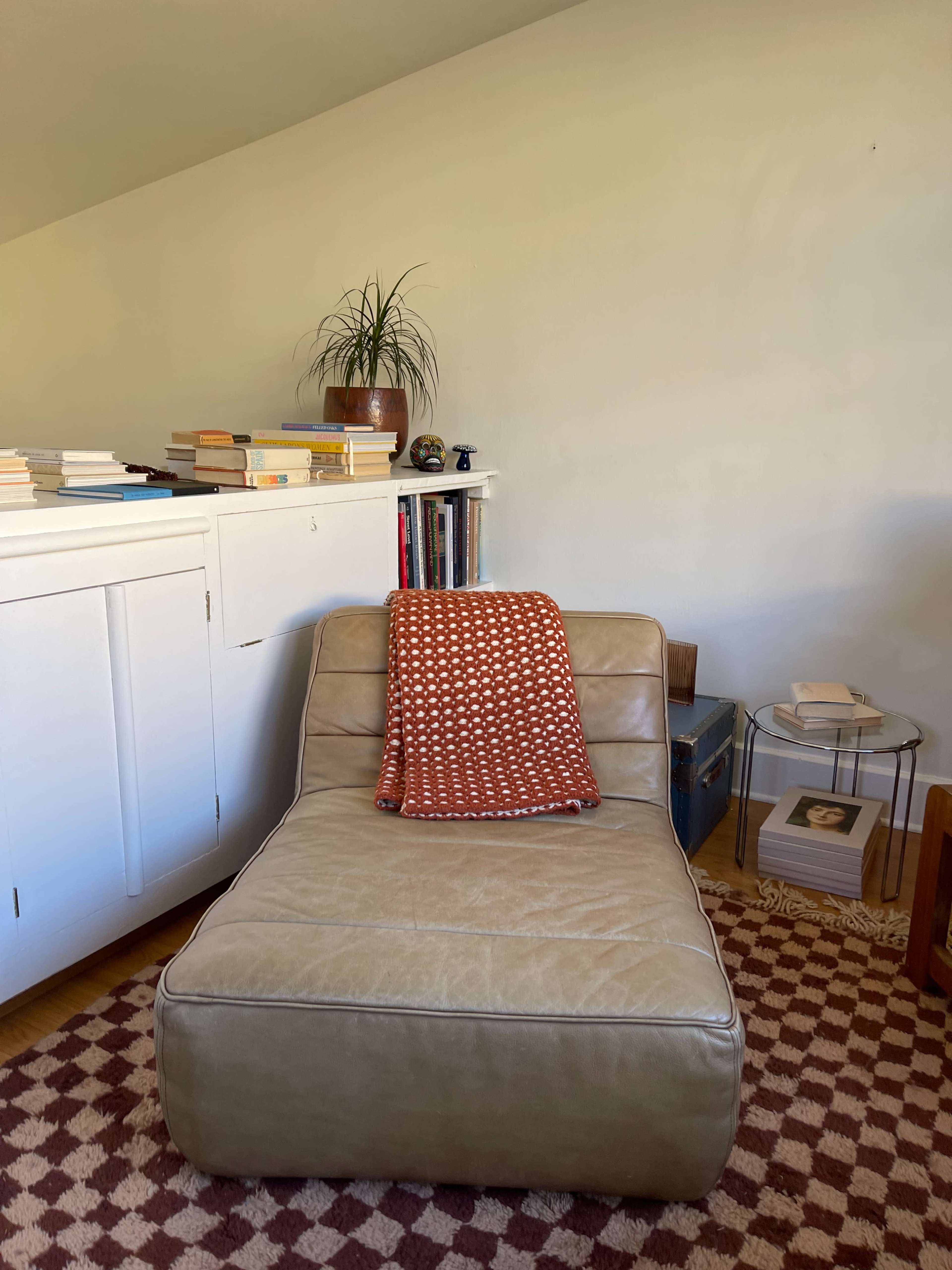 A light-colored chaise lounge with a patterned blanket rests beside a small side table and a bookshelf in a sunlit room.
