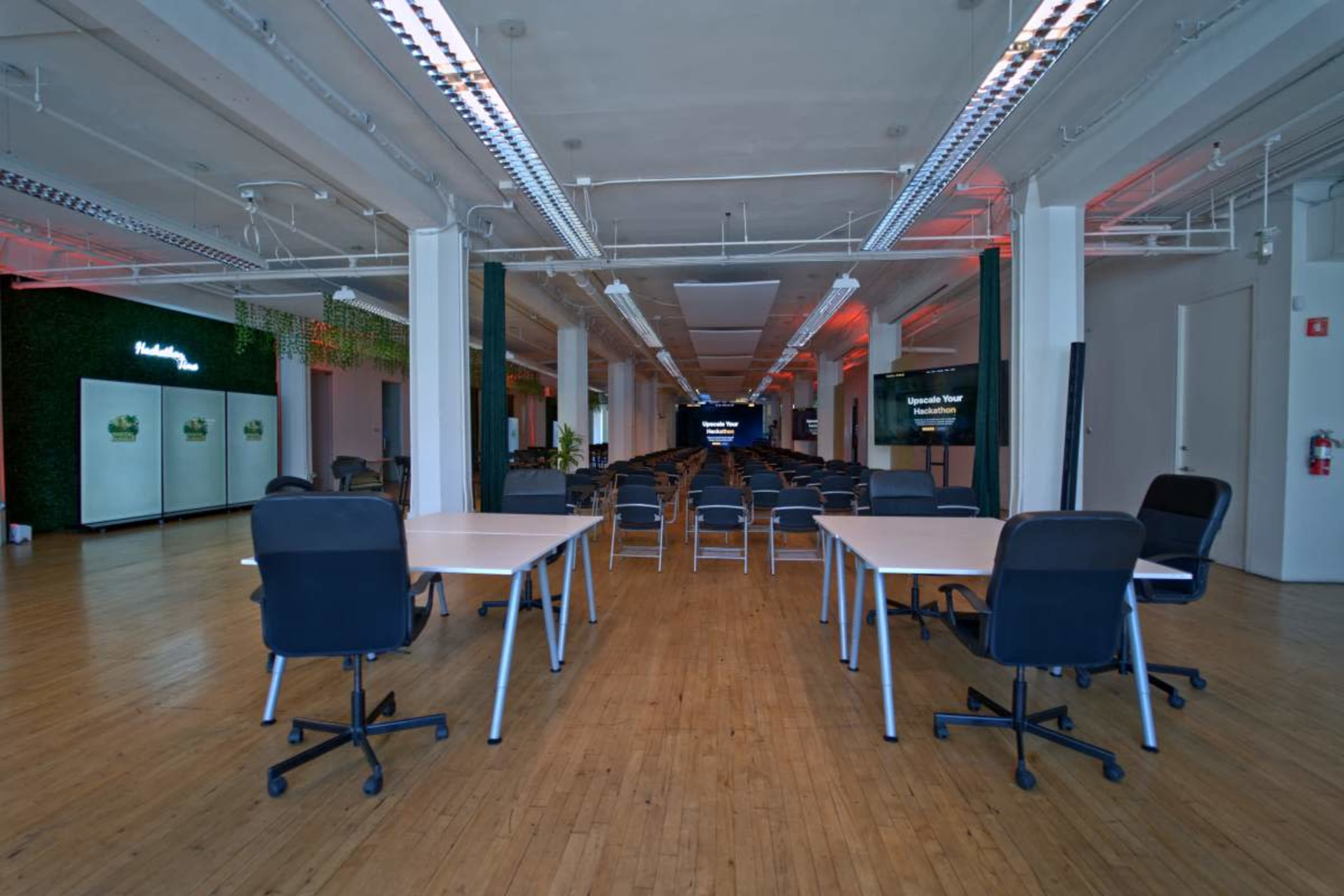 The image shows a spacious modern office with multiple empty desks arranged in rows, flanked by large screens displaying information.