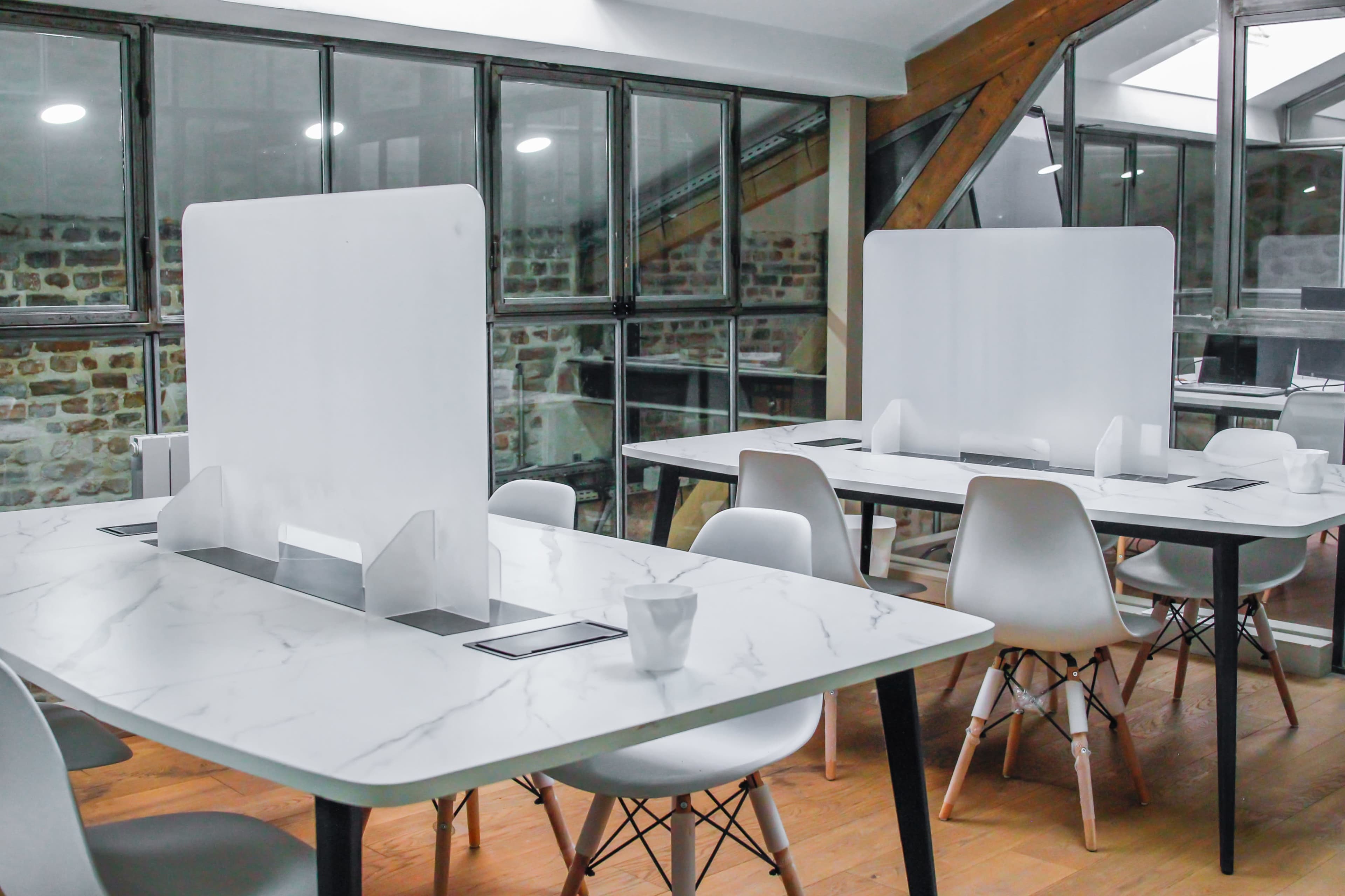 A modern workspace with several tables featuring marble tops and transparent dividers, surrounded by white chairs, all set in a room with exposed brick walls and large windows.
