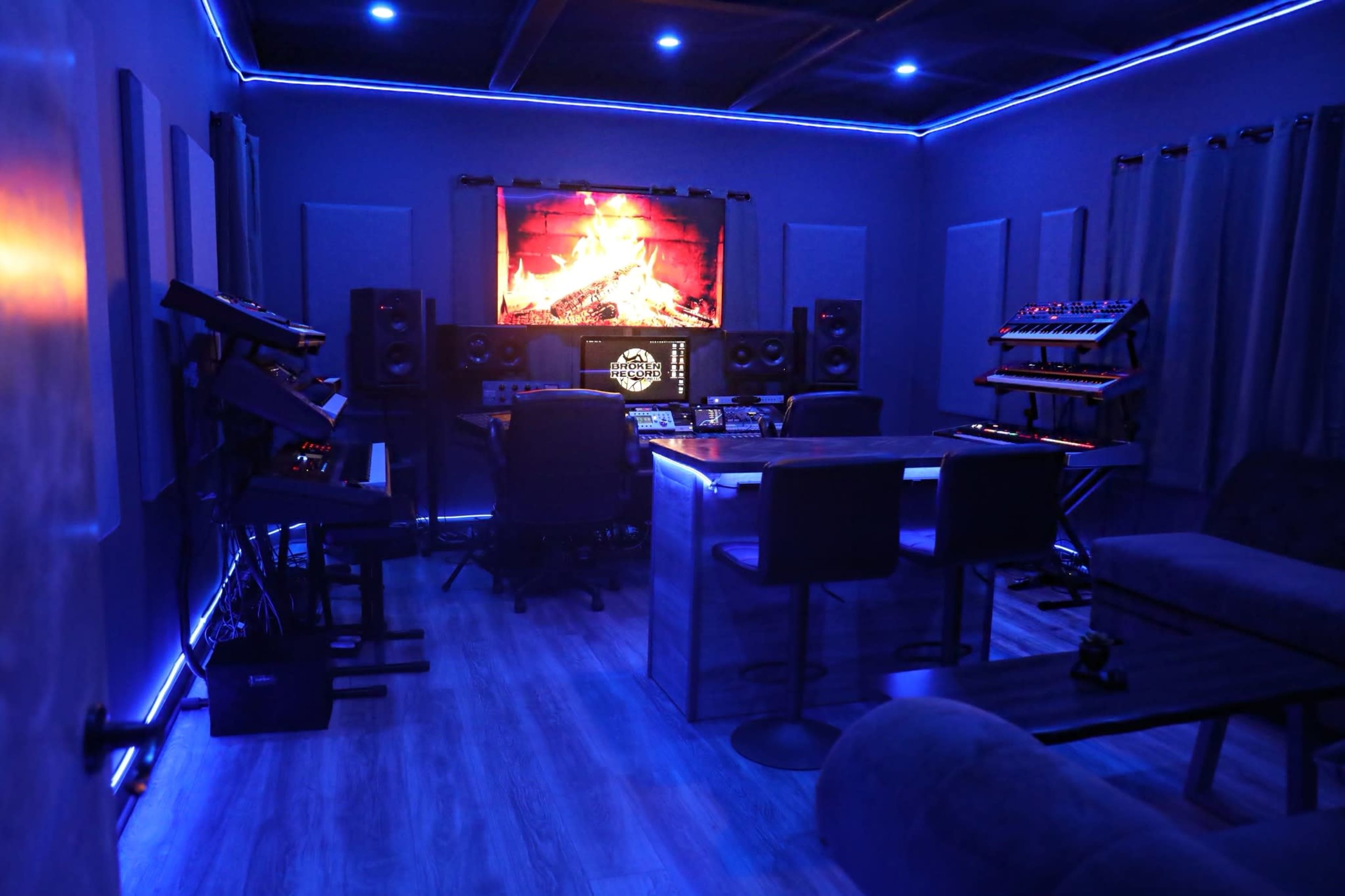A music production studio with blue walls, a large wall-mounted screen displaying a fireplace, and various electronic instruments arranged around a central table.