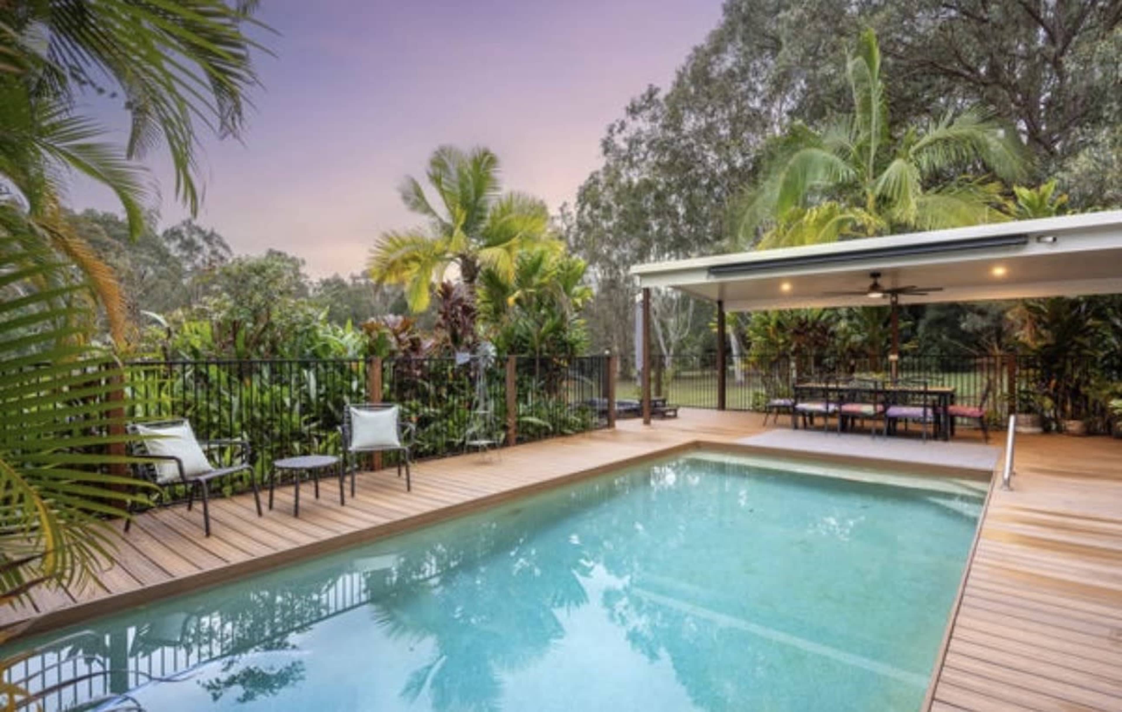 The image shows a modern swimming pool surrounded by a wooden deck, with tropical plants and a covered seating area nearby.