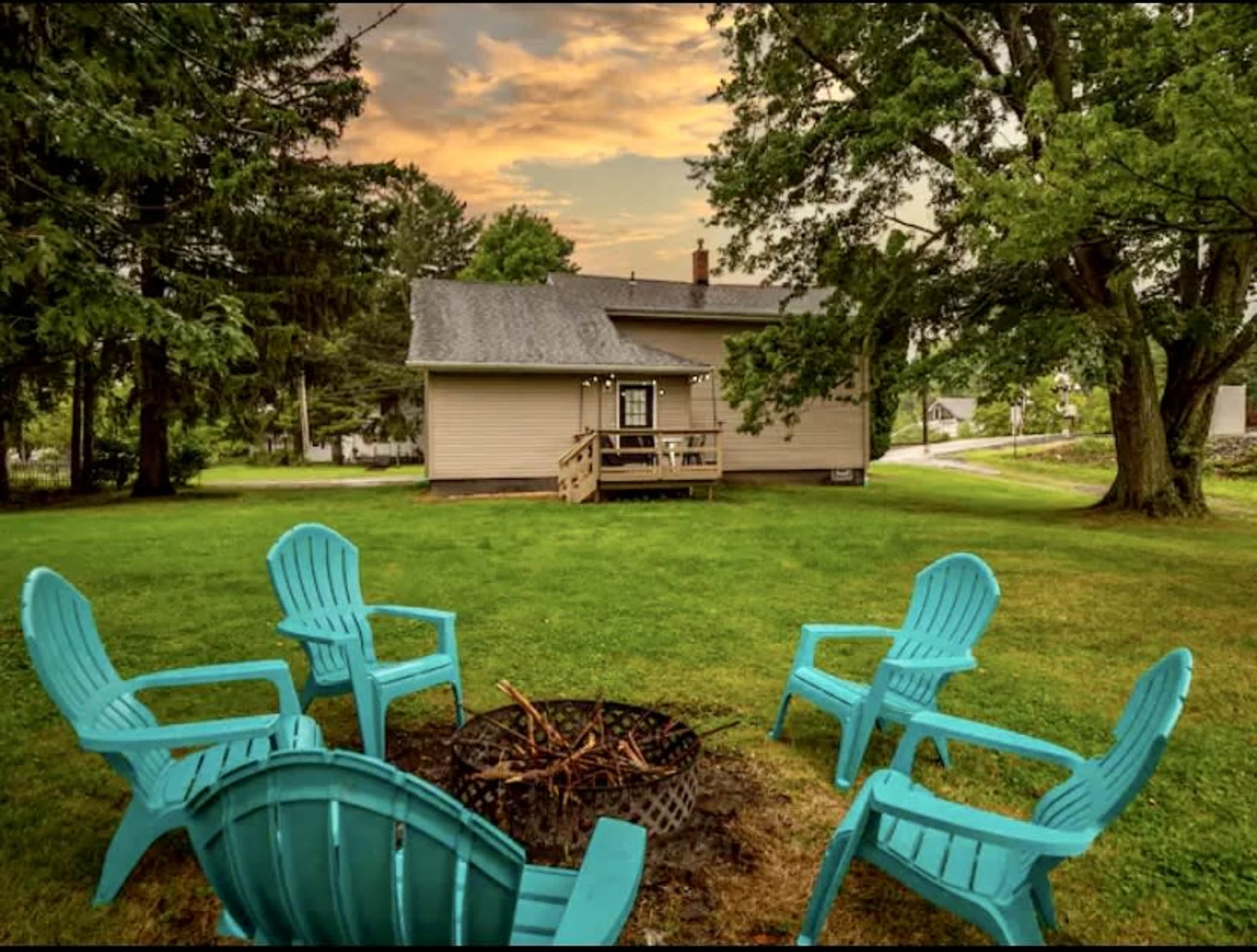 A fire pit surrounded by turquoise chairs is set in the grassy yard of a single-story house under a cloudy sky.