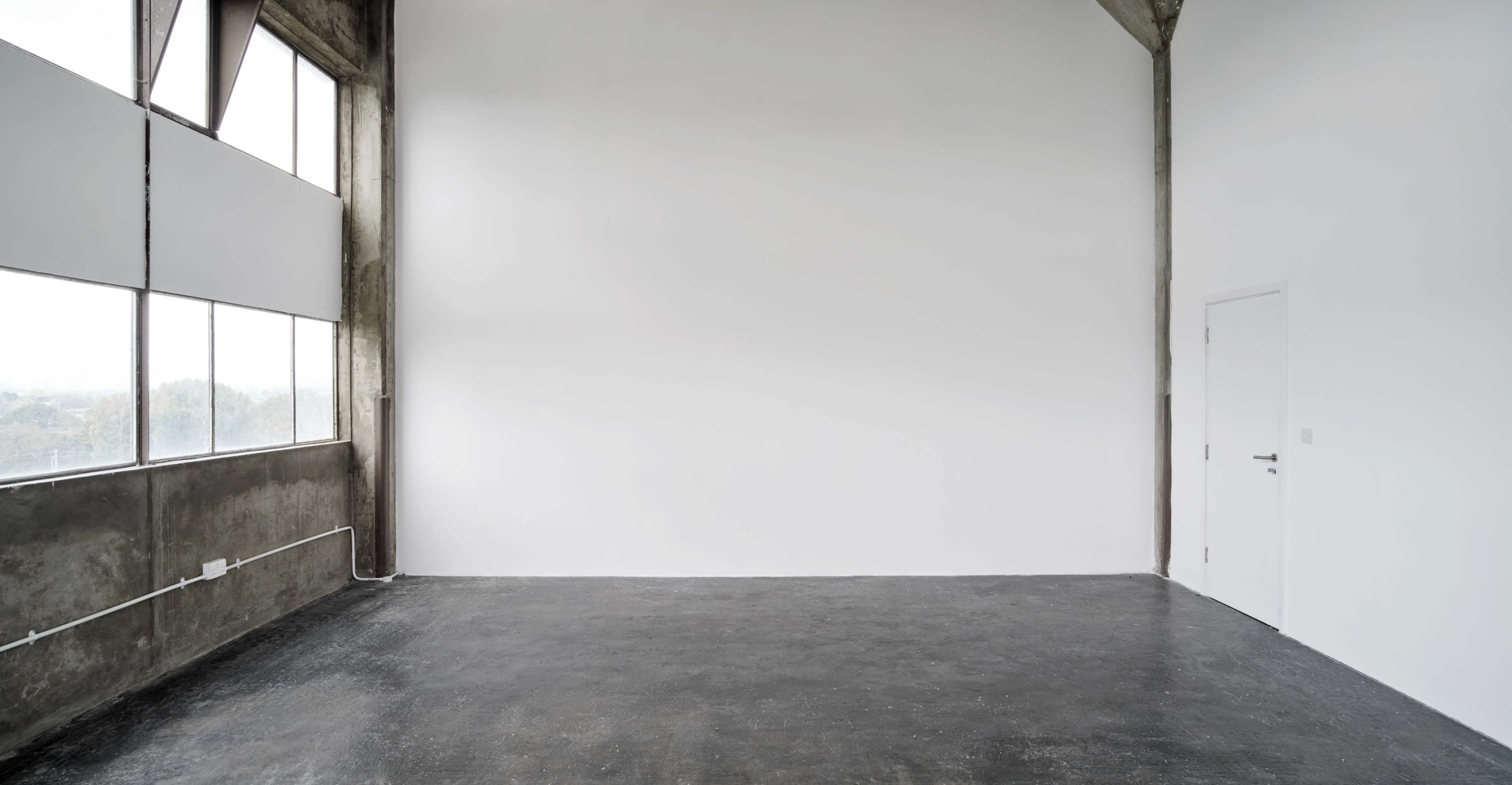 The image depicts a large, empty room with a concrete floor, tall windows on one side, and a blank wall opposite a door.