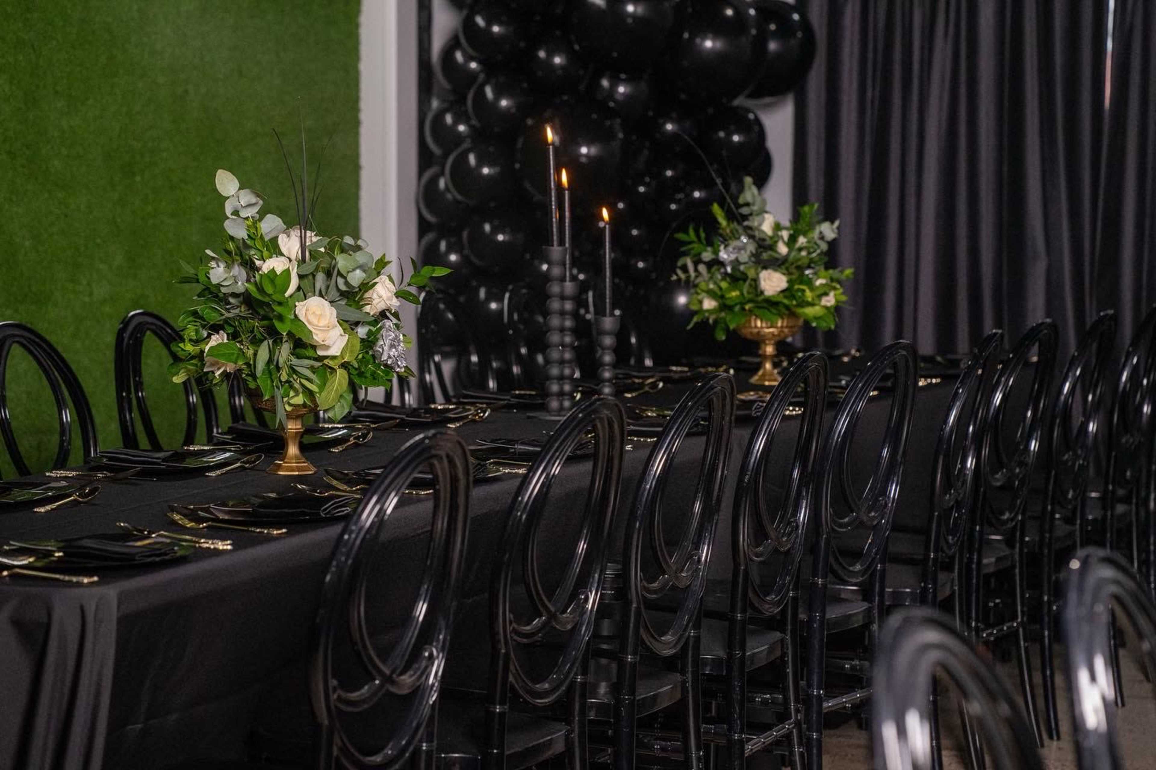 A long table is set with black tableware and floral arrangements, flanked by elegant black chairs, in a room featuring dark curtains and a greenery wall.